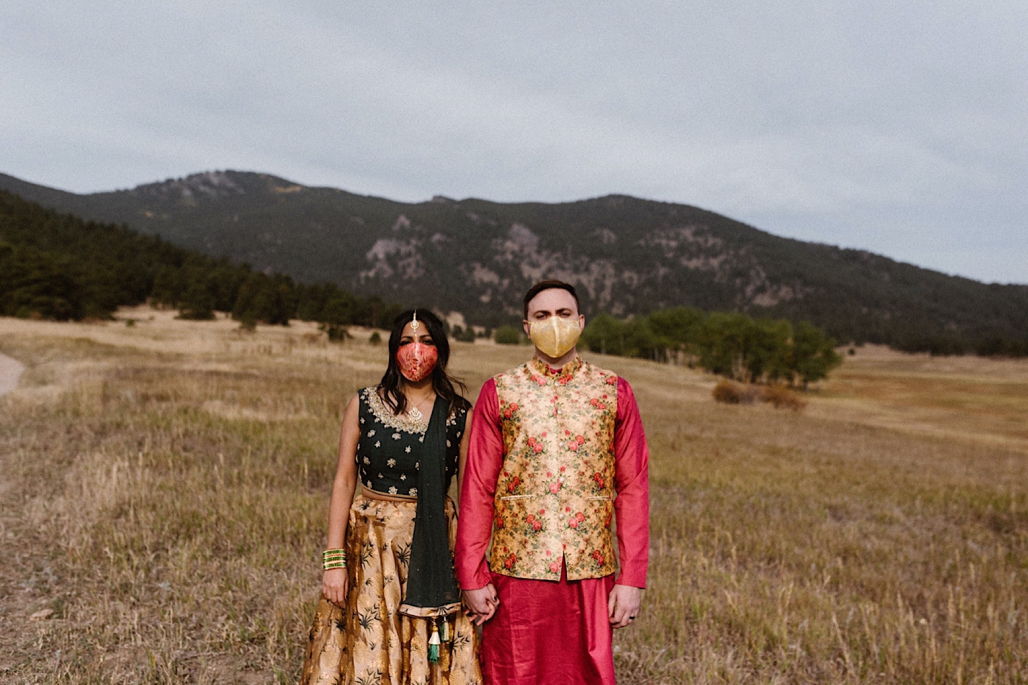 An Indian elopement couple stands in a field with mountains in the background as they stand side by side while holding hands with face masks covering their face. Photo by Denver elopement photographer, Colorado elopement photographer, Ashley Joyce Photography.
