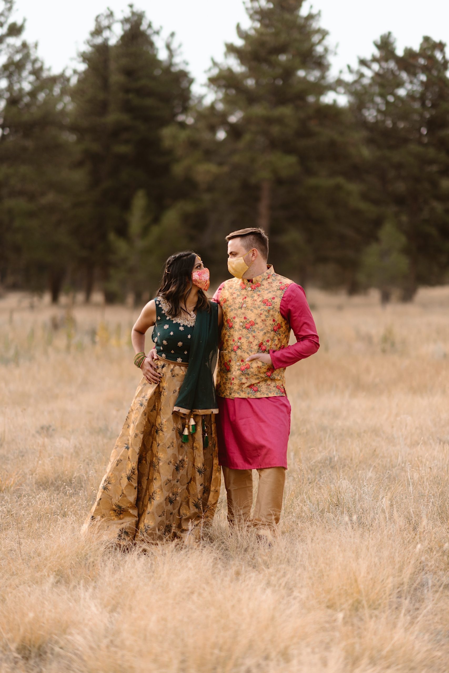 An Indian elopement couple stands in a field with mountains in the background as they stand side by side while holding hands with face masks covering their face. Photo by Denver elopement photographer, Colorado elopement photographer, Ashley Joyce Photography.