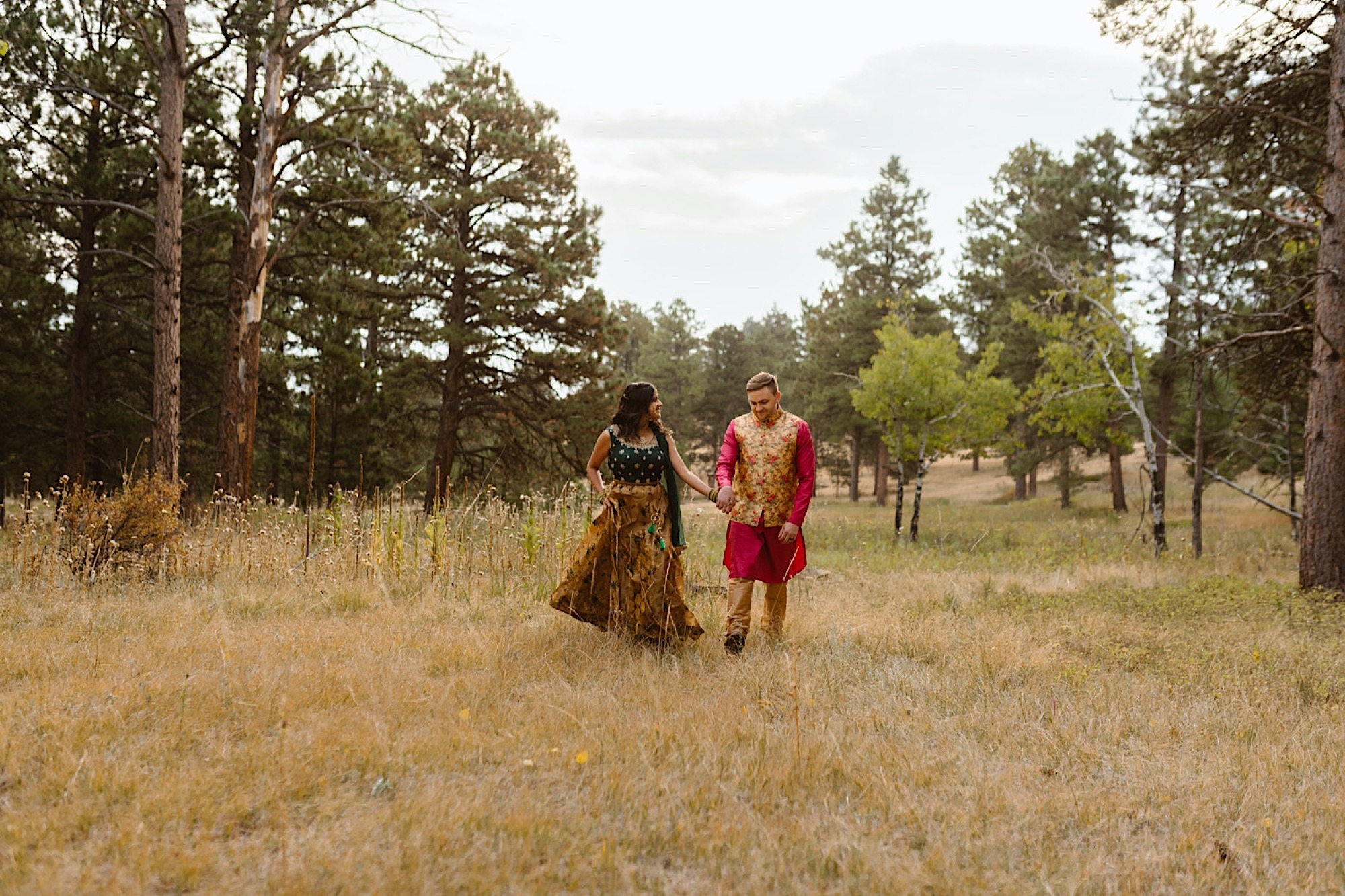 An Indian wedding couple stands in a field of evergreen trees near Denver, Colorado as they walk together for their Indian elopement photos. Photo by Denver elopement photographer, Colorado elopement photographer, Ashley Joyce Photography.