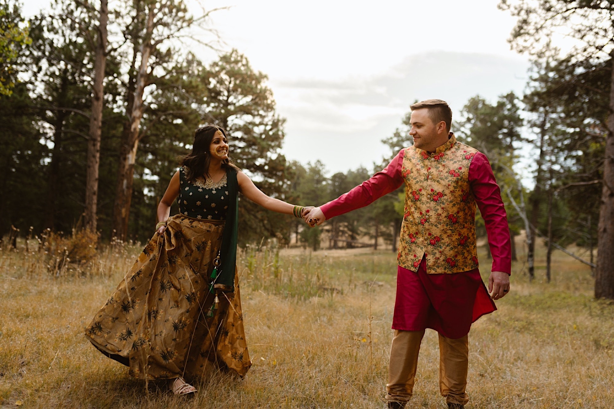 An Indian wedding couple stands in a field of evergreen trees near Denver, Colorado as they walk together for their Indian elopement photos. Photo by Denver elopement photographer, Colorado elopement photographer, Ashley Joyce Photography.