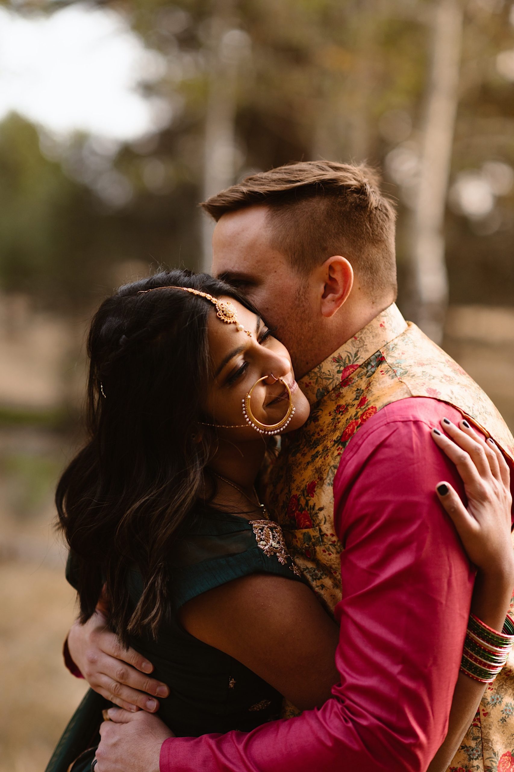 An Indian wedding couple stands in a field of evergreen trees near Denver, Colorado as they embrace one another for their Indian elopement photos. Photo by Denver elopement photographer, Colorado elopement photographer, Ashley Joyce Photography.