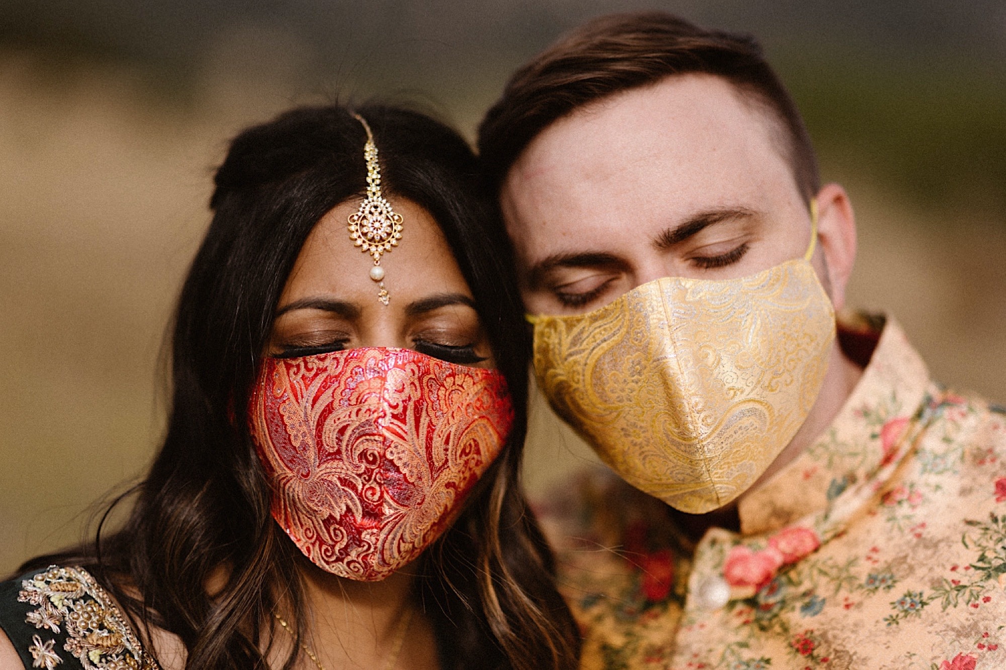 An Indian elopement couple stands in a field as their faces are covered in face mask for their pandemic elopement. They stand with their colorful masks as they close their eyes. Photo by Denver elopement photographer, Colorado elopement photographer, Ashley Joyce Photography.