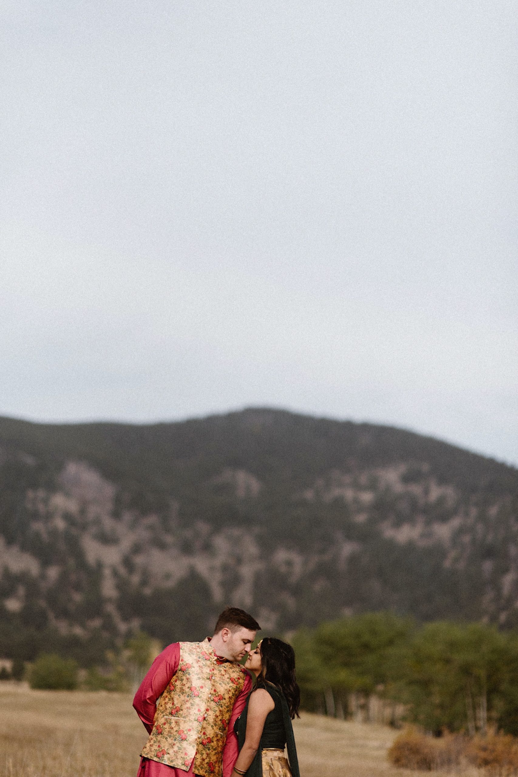 An Indian elopement couple stands in a field with mountains in the background as they stand side by side while holding hands with face masks covering their face. Photo by Denver elopement photographer, Colorado elopement photographer, Ashley Joyce Photography.