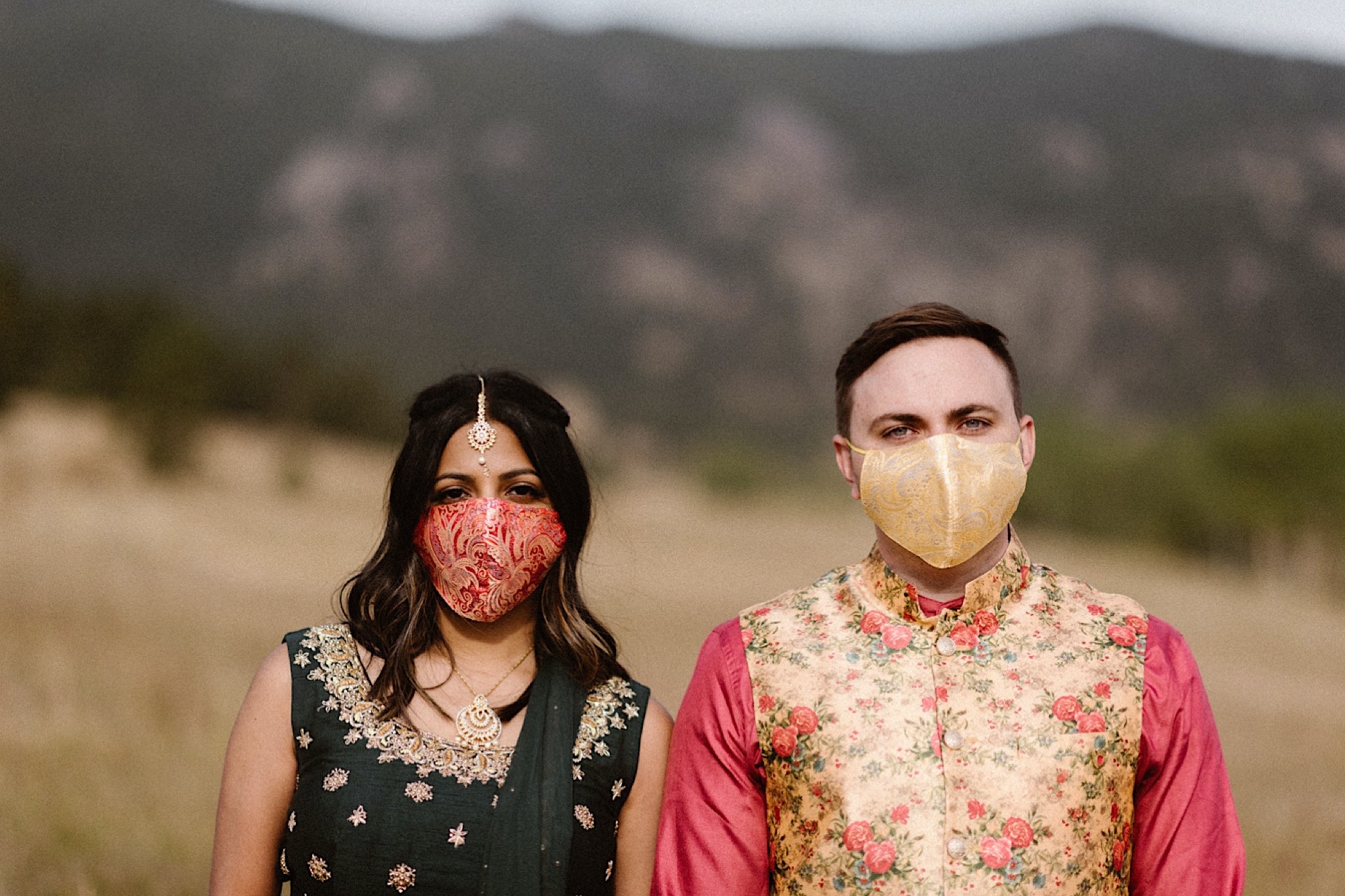 An Indian elopement couple stands in a field with mountains in the background as they stand side by side while holding hands with face masks covering their face. Photo by Denver elopement photographer, Colorado elopement photographer, Ashley Joyce Photography.