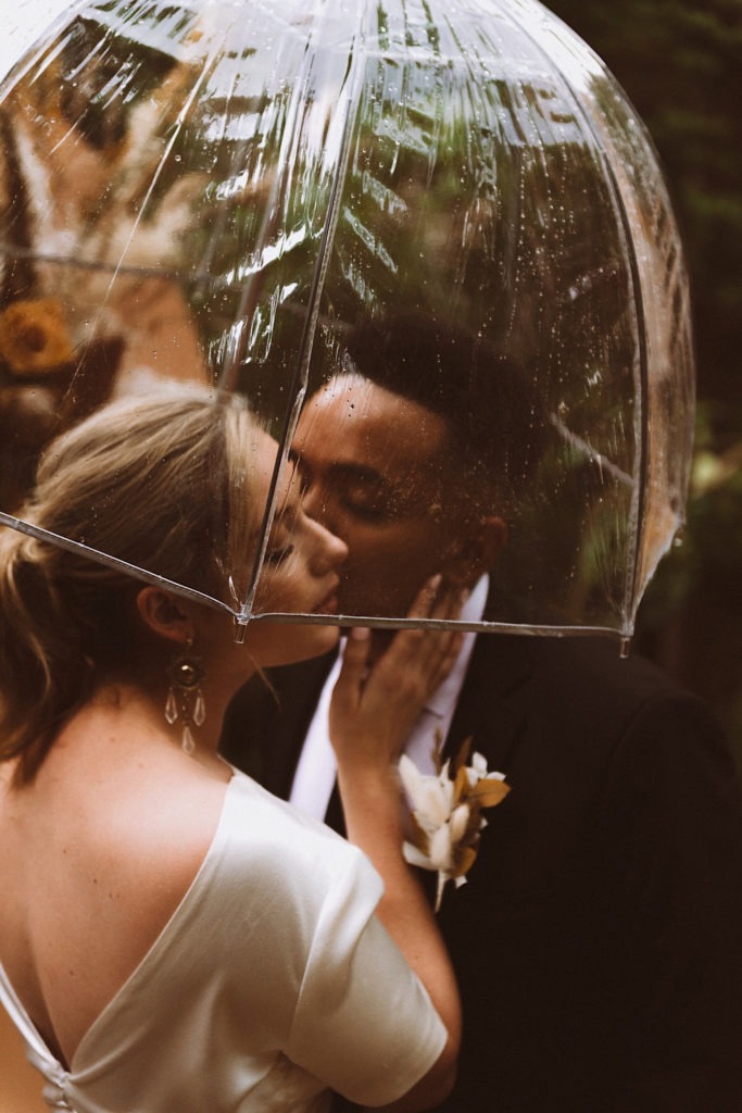 Groom kissing bride on the cheek under a clear umbrella | Wedding portraits | Elopement portraits | Bride and groom elopement photos | Elopement wedding photos in Colorado | Colorado wedding photos | Colorado elopement photos | Elopement at Treehaus, Colorado | Treehaus Colorado | Colorado elopement | Treehaus elopement | Colorado elopement venue | Colorado mountain elopement venue | Unique elopement venue | Elopement venue in Colorado | Ashley Joyce Photography 2020