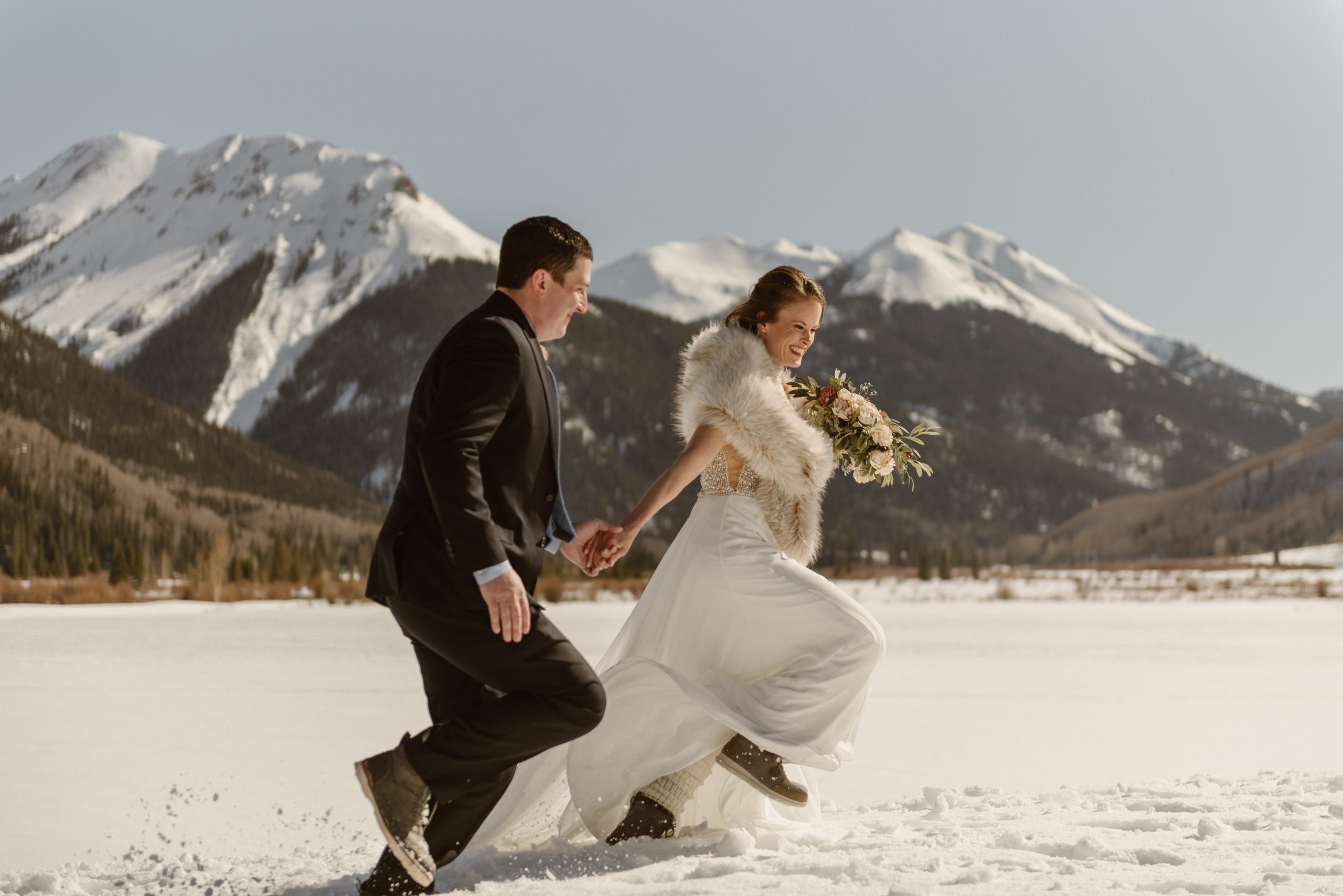 A Ouray elopement couple runs through the snow in their wedding attire with mountains in the background. Photo by Denver elopement photographer, Colorado elopement photographer, Ashley Joyce Photography.