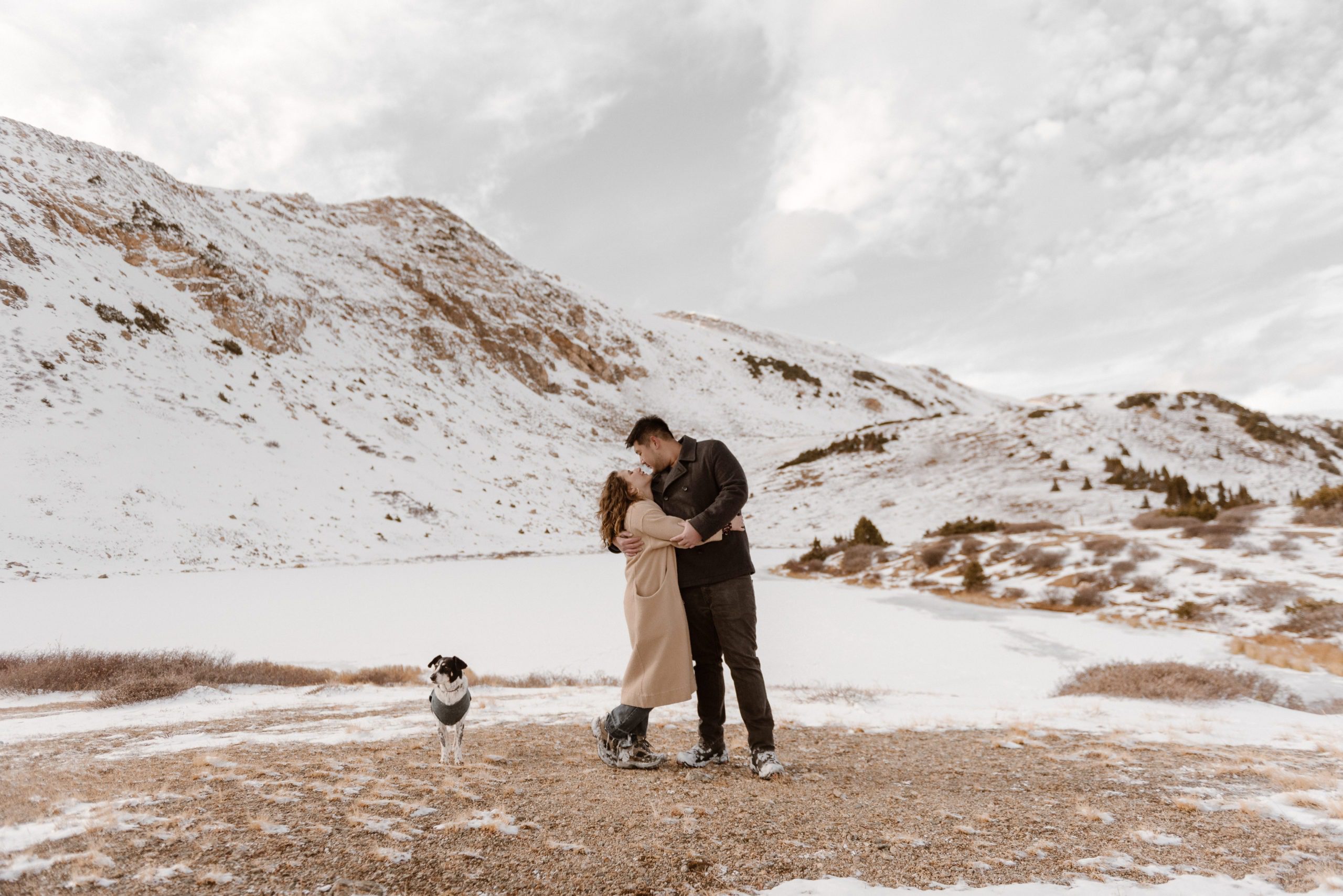 A Colorado couple stands on the snowy ground with their dog for their Loveland Pass couples session. Photo by Denver elopement photographer, Ashley Joyce Photography. Copyright 2022.