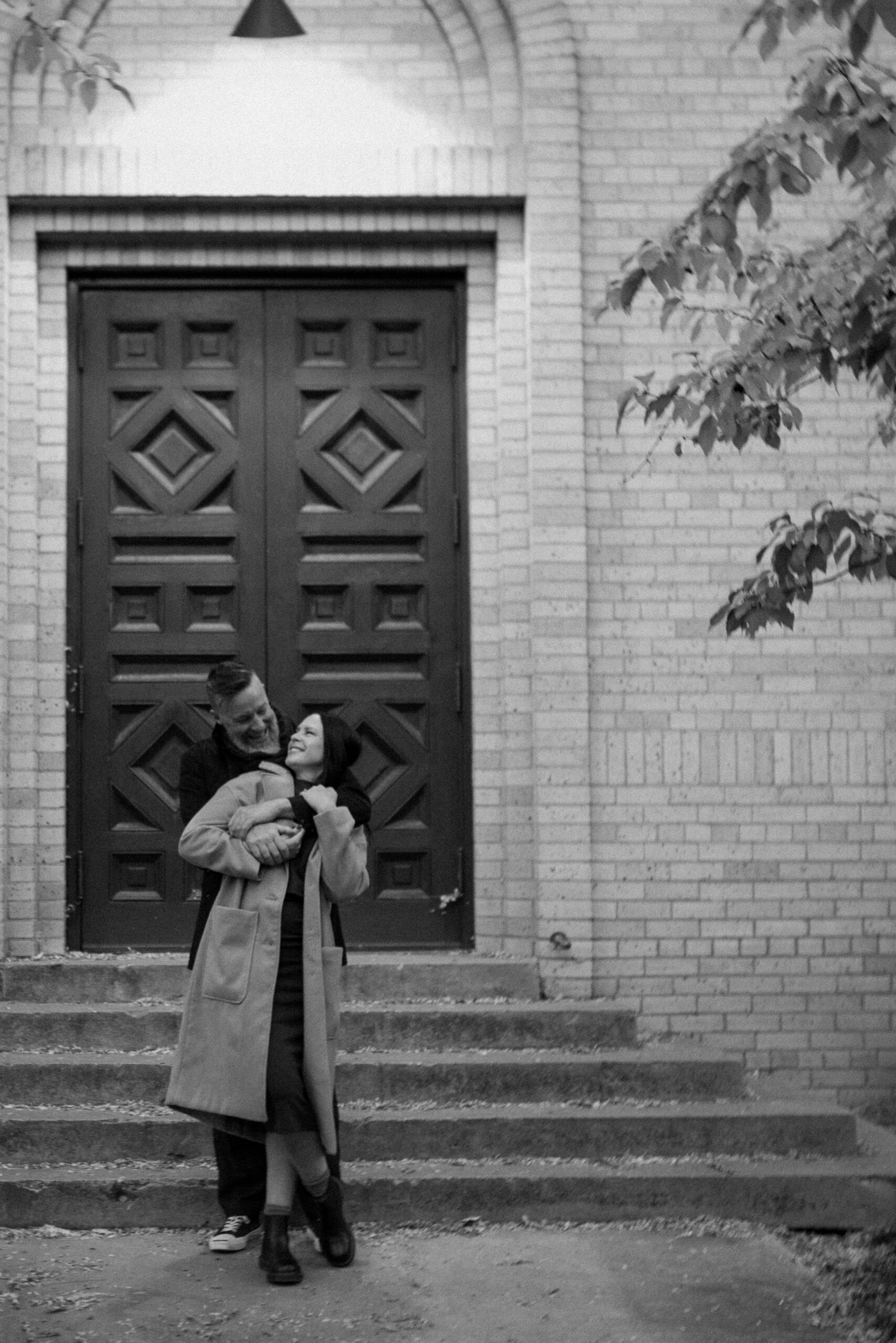A black and white photo of an engaged couple embracing one another in front of The Smiley Building in Durango, Colorado for their engagement session, taken by Colorado wedding photographer, Ashley Joyce.