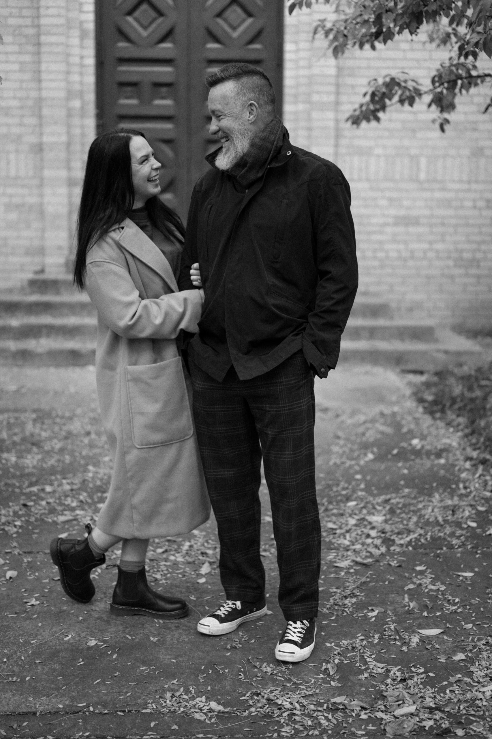 A black and white photo of an engaged couple holding hands while they laugh in front of The Smiley Building in Durango, Colorado for their engagement session, taken by Colorado wedding photographer, Ashley Joyce.