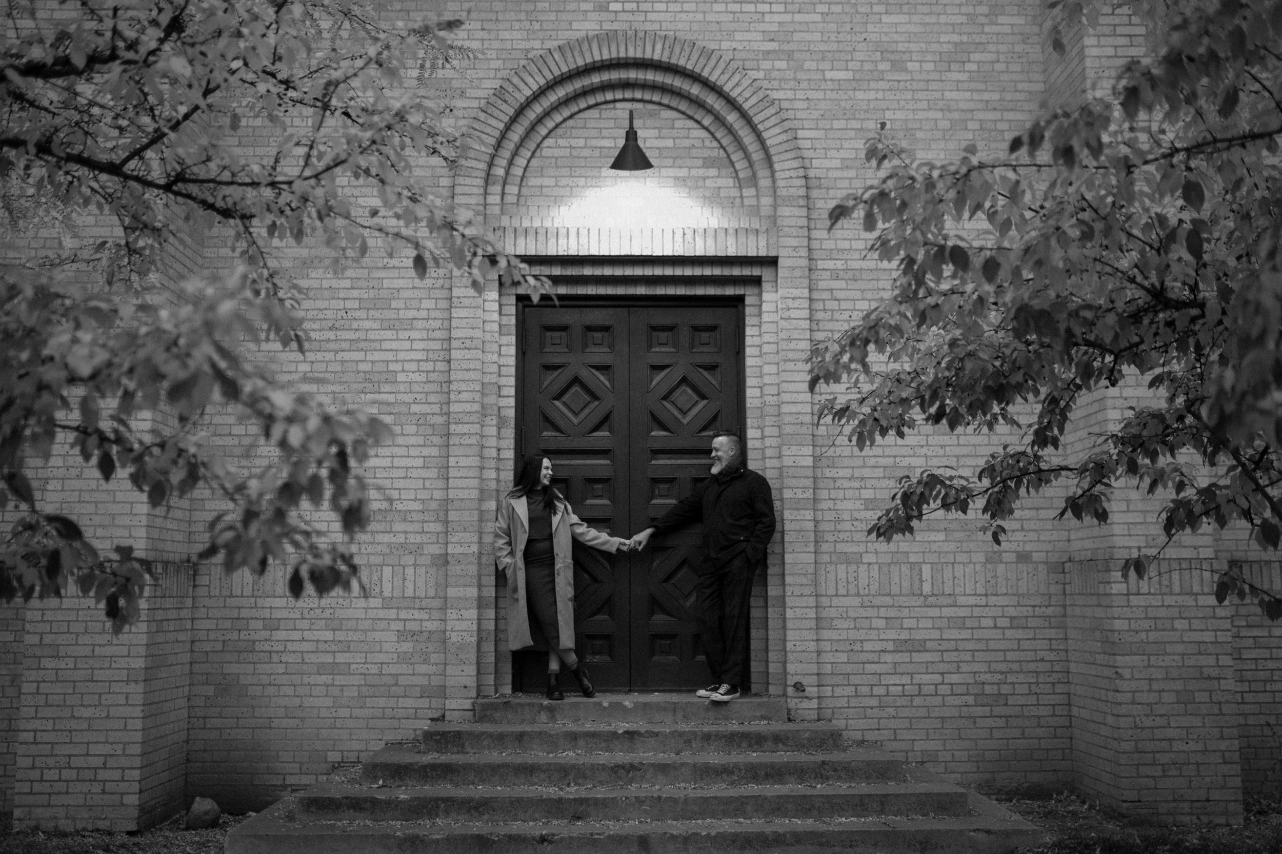 A black and white photo of an engaged couple holding hands in front of The Smiley Building in Durango, Colorado for their engagement session, taken by Colorado wedding photographer, Ashley Joyce.