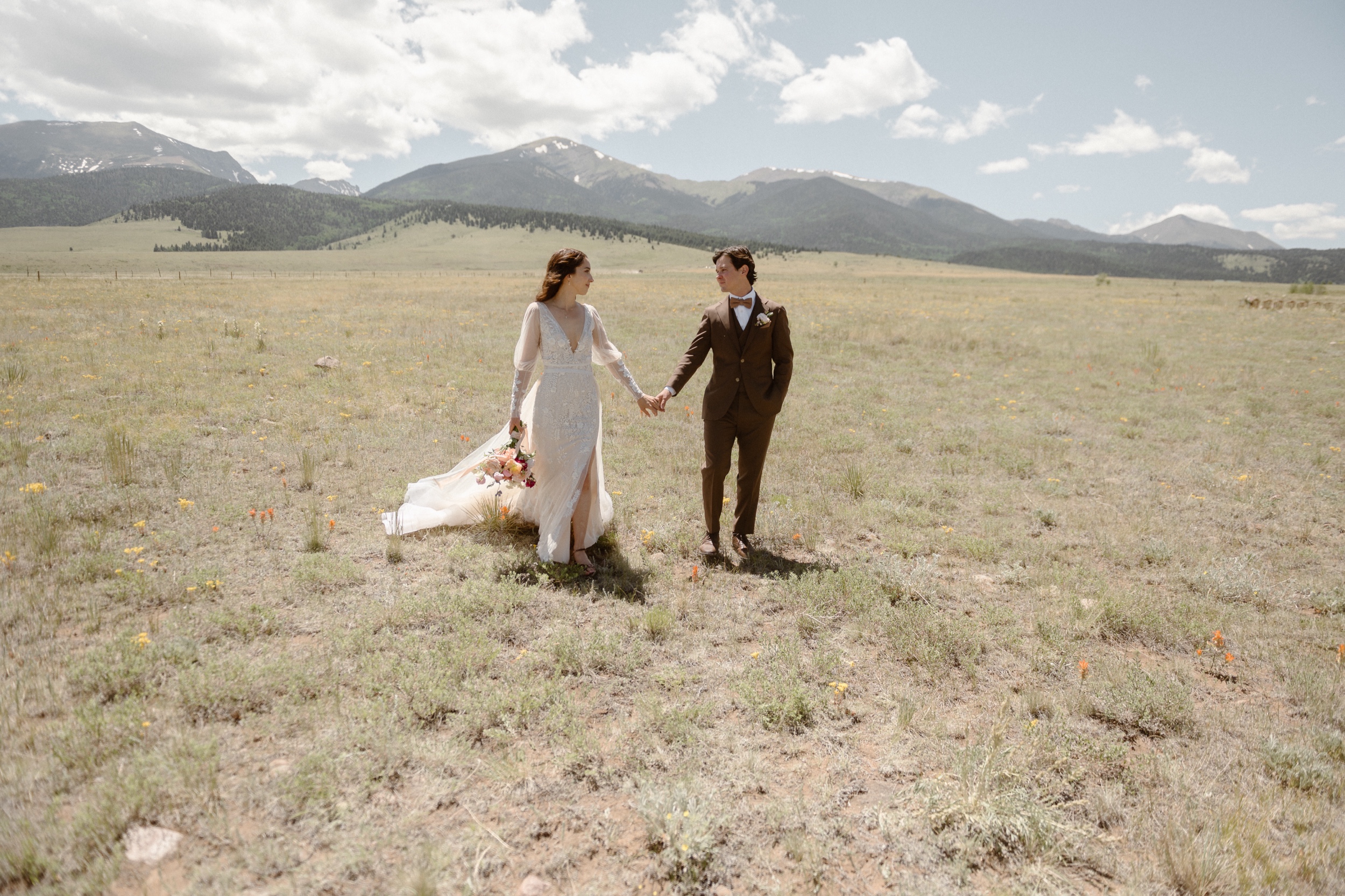 A bride and groom walk towards the camera while holding hands in a grassy field with mountains in the background at Three Peaks Ranch. Photo by Colorado wedding photographer Ashley Joyce.