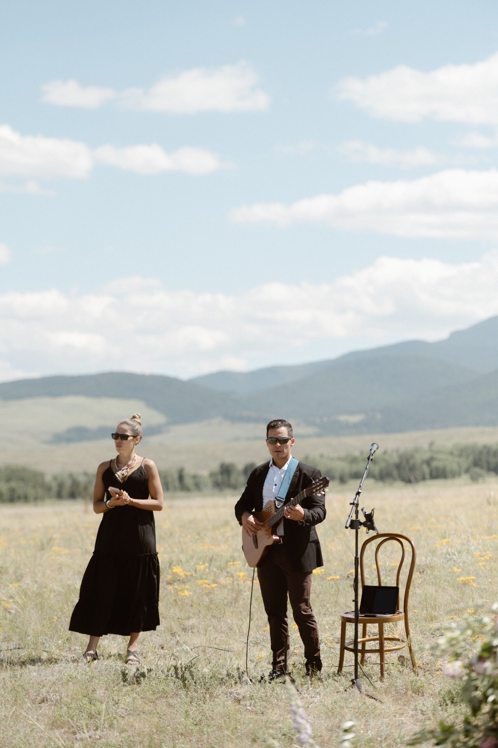 Two musicians stand in a grassy field with mountains in the background. Photo by Colorado wedding photographer Ashley Joyce.