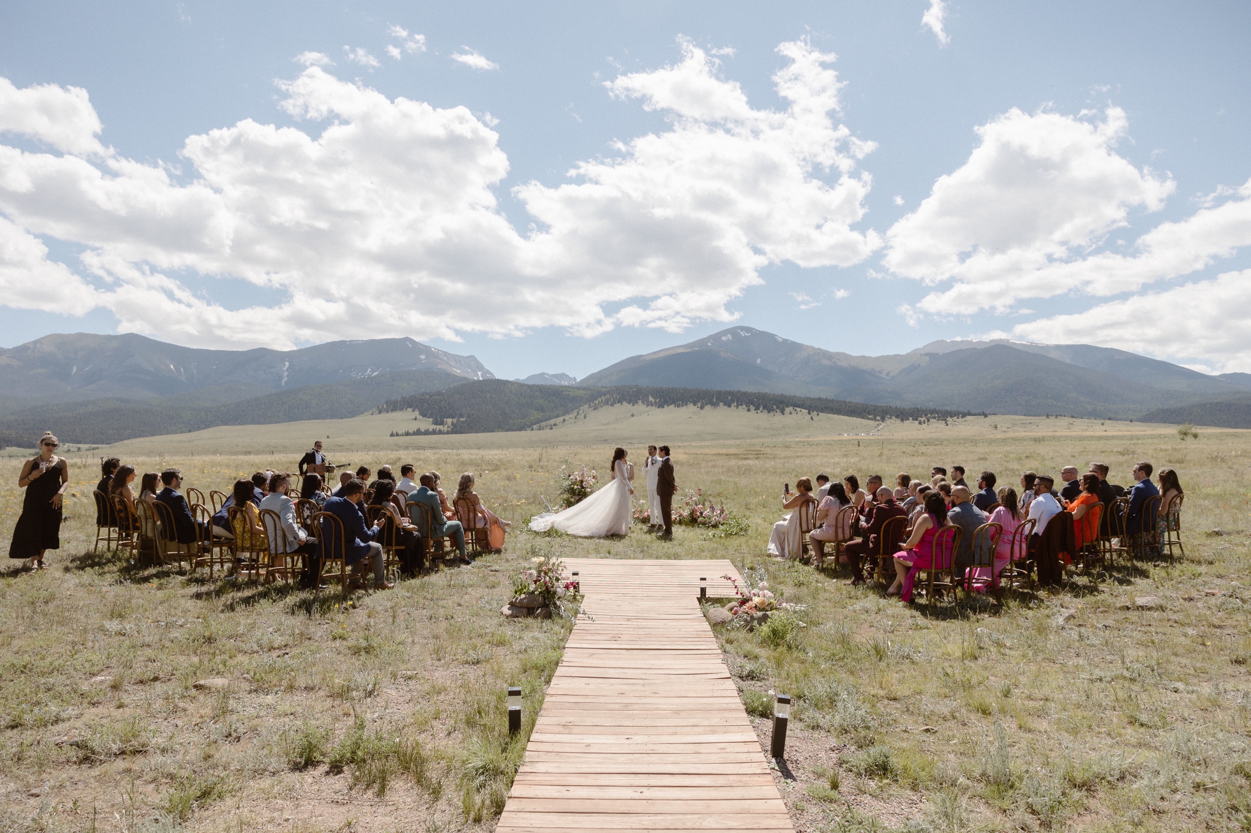 A wide view of a bride and groom standing in a grassy field with mountains in the background as they host their Three Peaks Ranch wedding. Photo by Colorado wedding photographer Ashley Joyce.