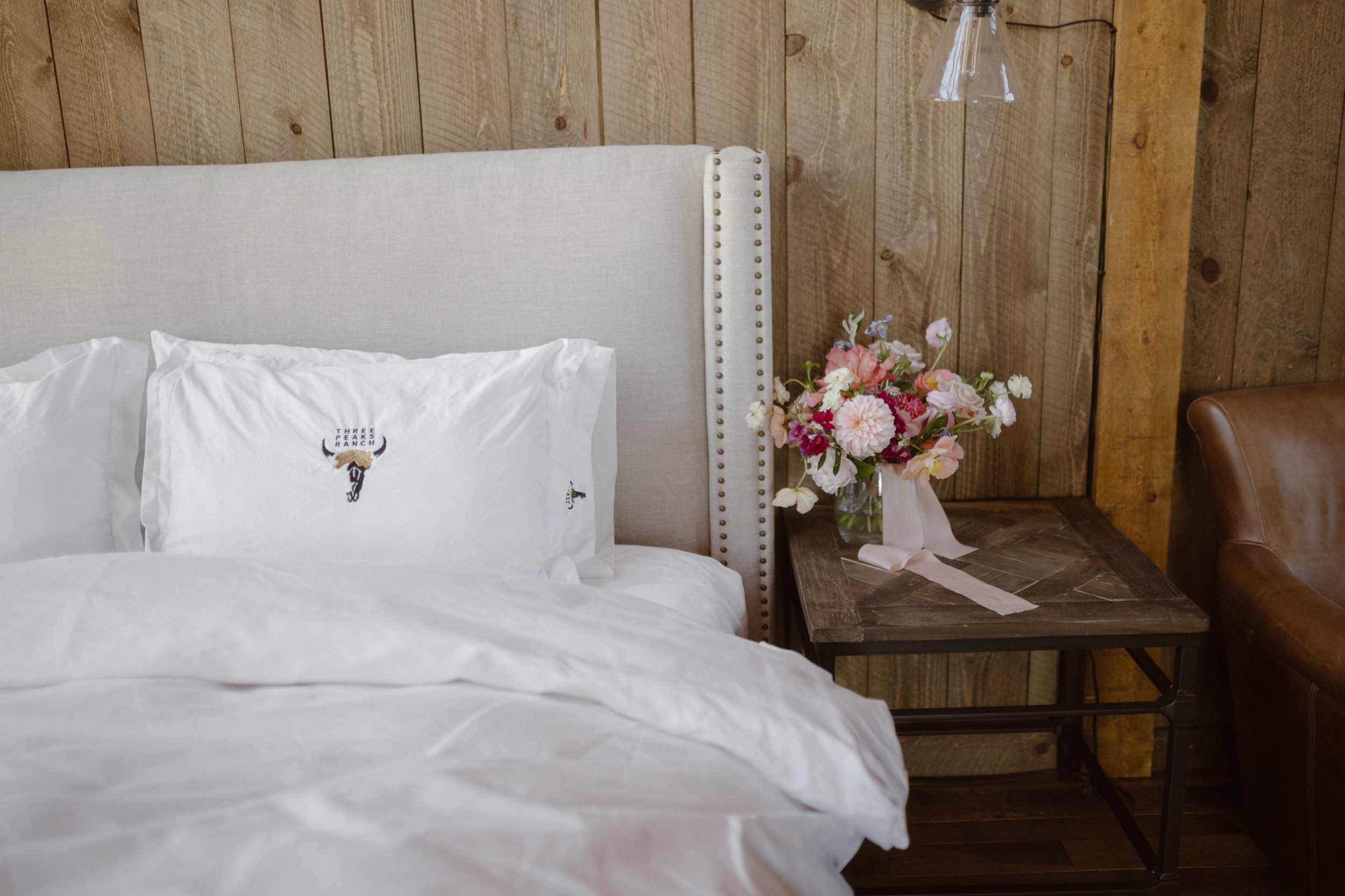 A photo of a wedding bouquet on a bedside table next to a white linen-covered bed at Three Peaks Ranch. Photo by Ashley Joyce.