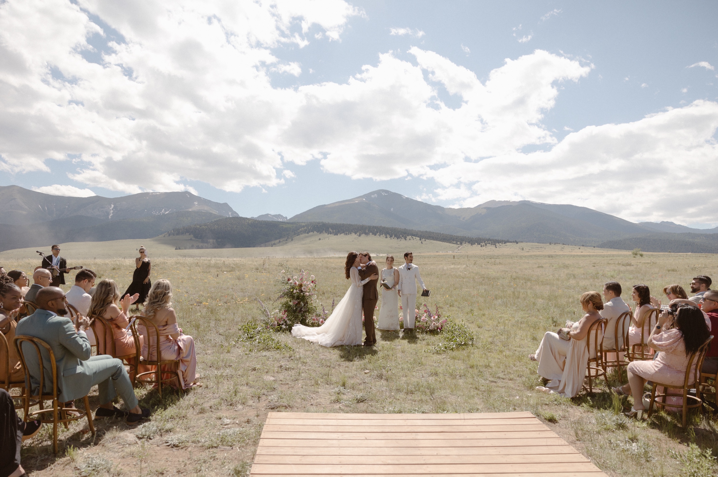 A bride and groom kiss one another after their wedding ceremony in a grassy field with mountains in the background. Photo by Ashley Joyce.