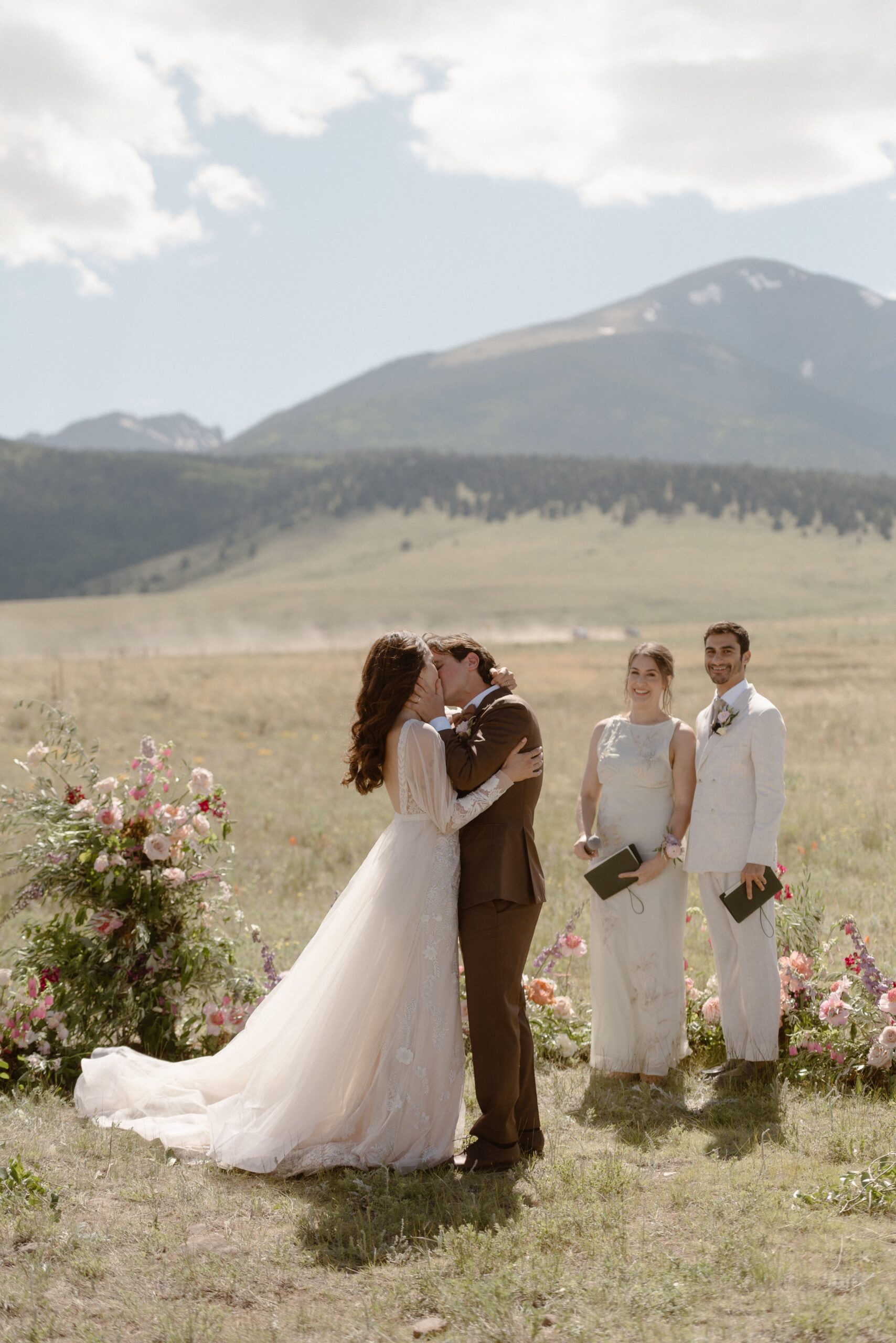 A bride and groom kiss one another after their wedding ceremony in a grassy field with mountains in the background. Photo by Ashley Joyce.