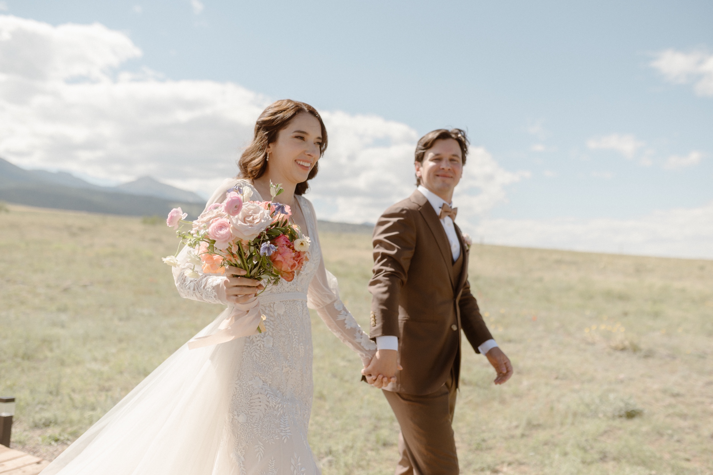 A bride and groom walk along a grassy field as they hold hands. Photo by Colorado wedding photographer Ashley Joyce.