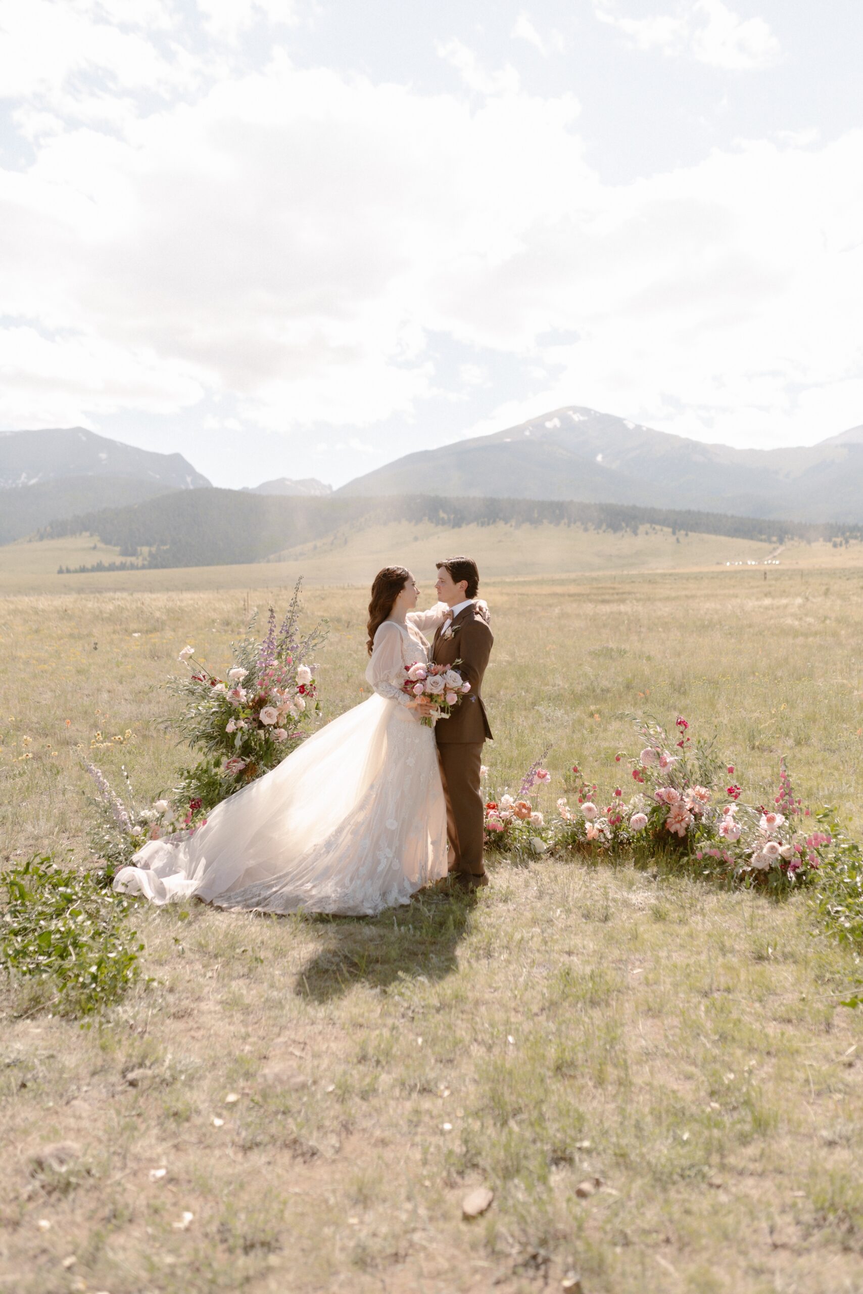 A bride and groom pose for their wedding portraits at their Three Peaks ranch wedding in a grassy field with mountains in the background. Photo by Colorado wedding photographer Ashley Joyce.