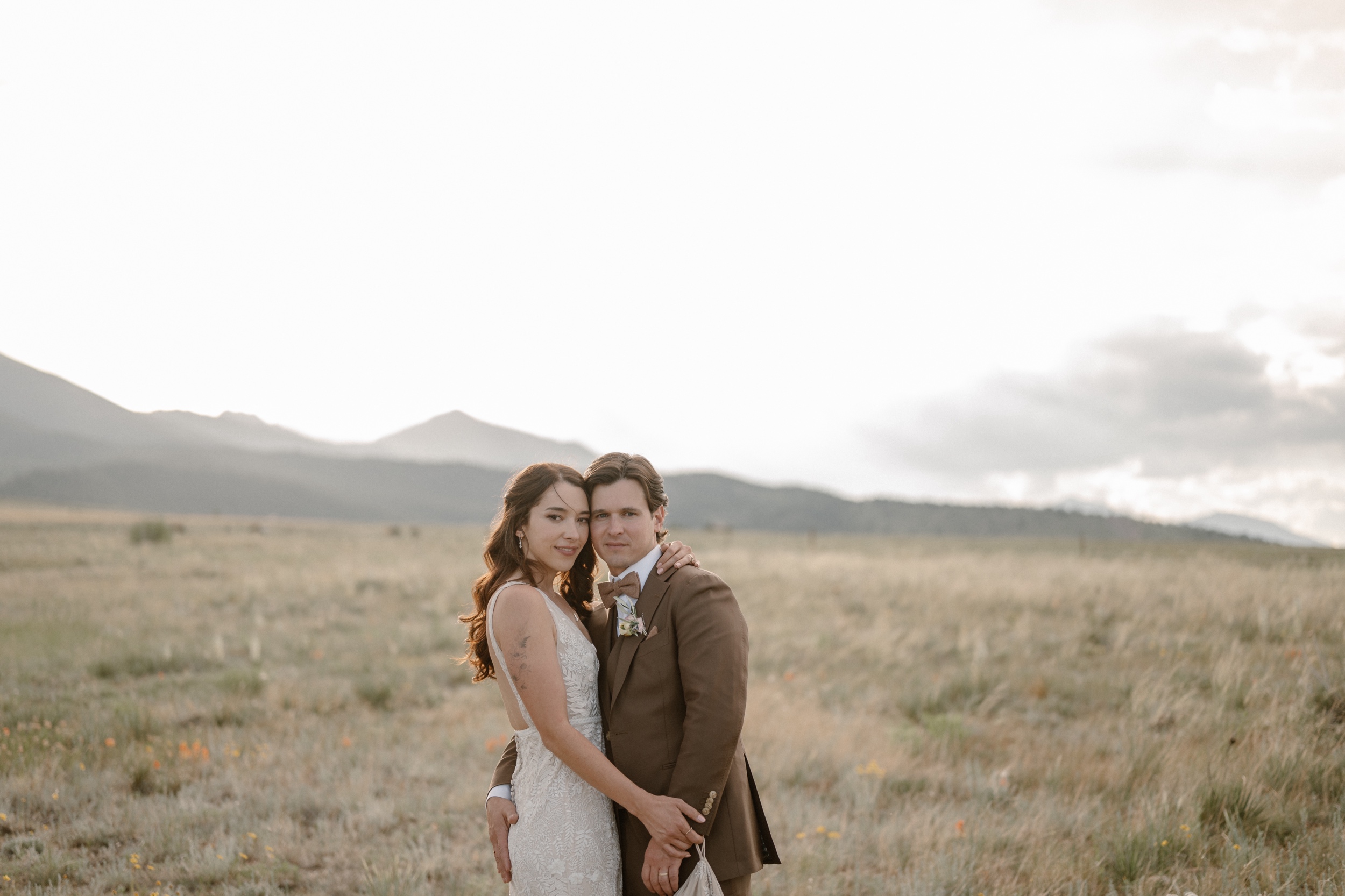 A bride and groom pose for their wedding portraits at their Three Peaks ranch wedding in a grassy field with mountains in the background. Photo by Colorado wedding photographer Ashley Joyce.