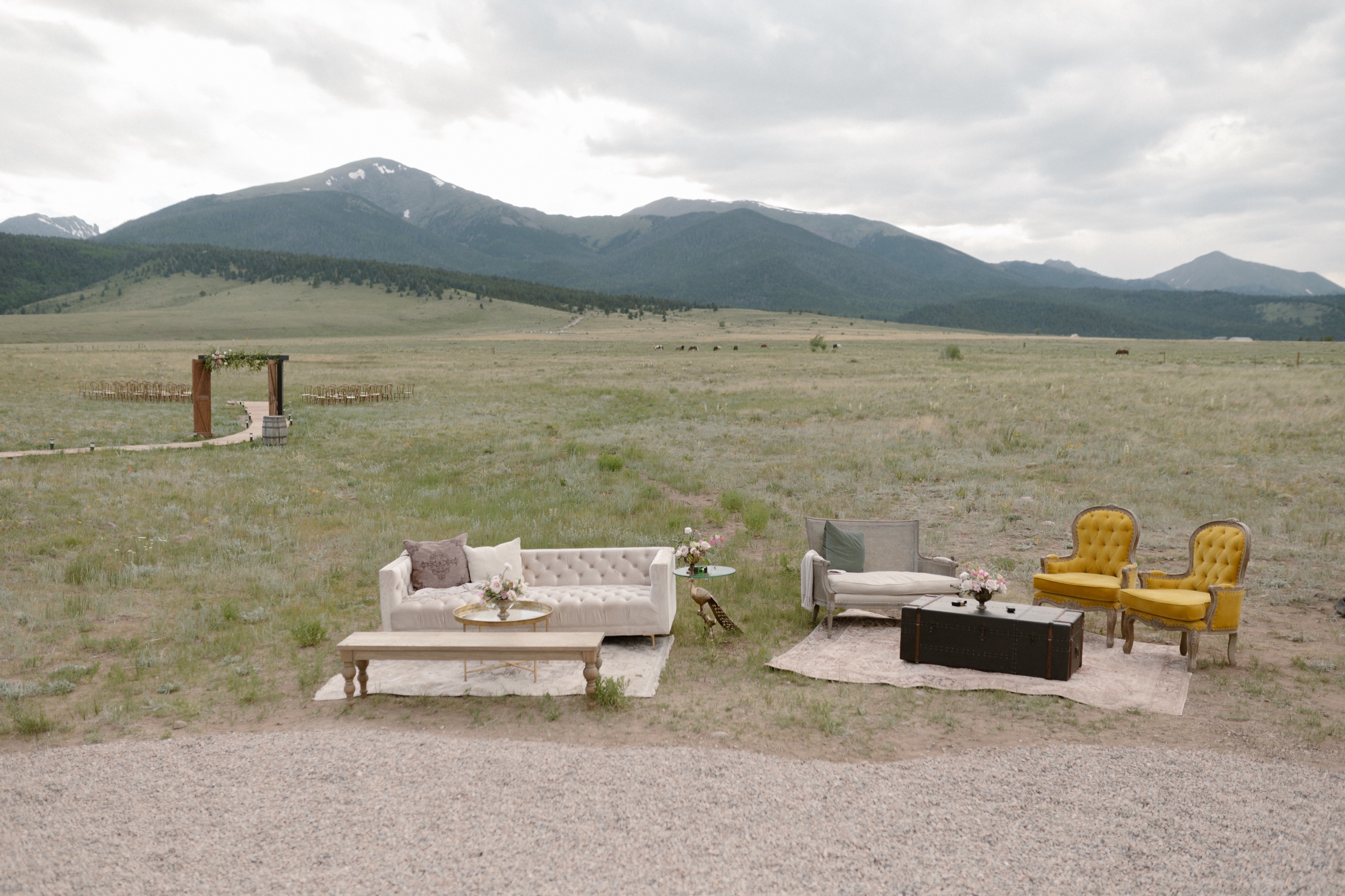 A photo of lounge decor on the grass with a minimal ceremony setup in the background with tall mountains in the far back. Photo taken at Three Peaks Ranch by Ashley Joyce.