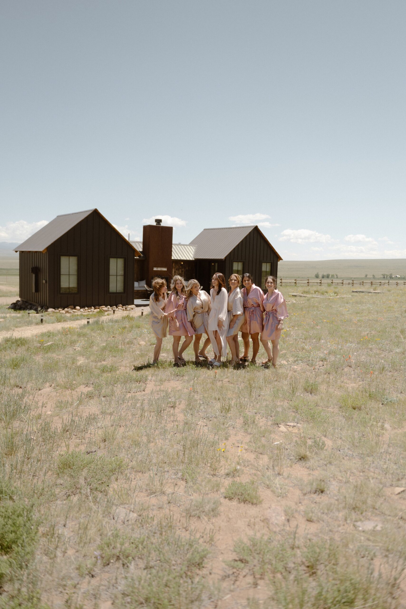 A bride and her bridesmaids and matronly family members stand in the grass field with a cabin in the background. Photo by Ashley Joyce.