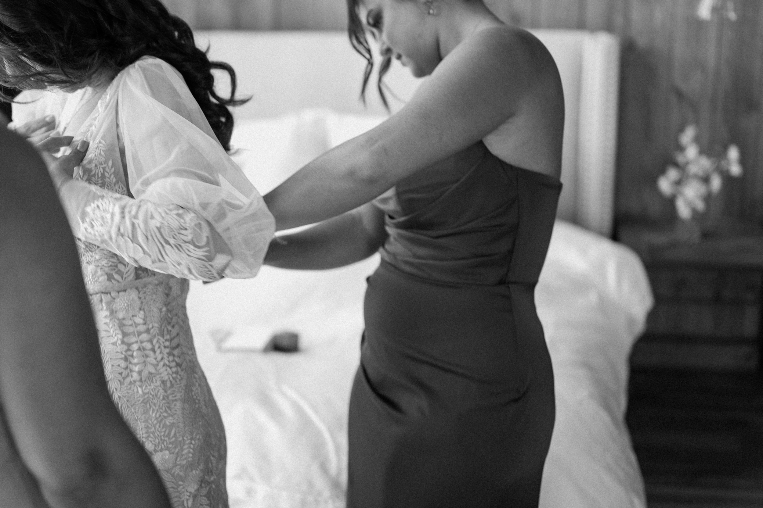 A black and white photo of a bridesmaid helping a bride get in her wedding dress at Three Peaks Ranch. Photo by Ashley Joyce.