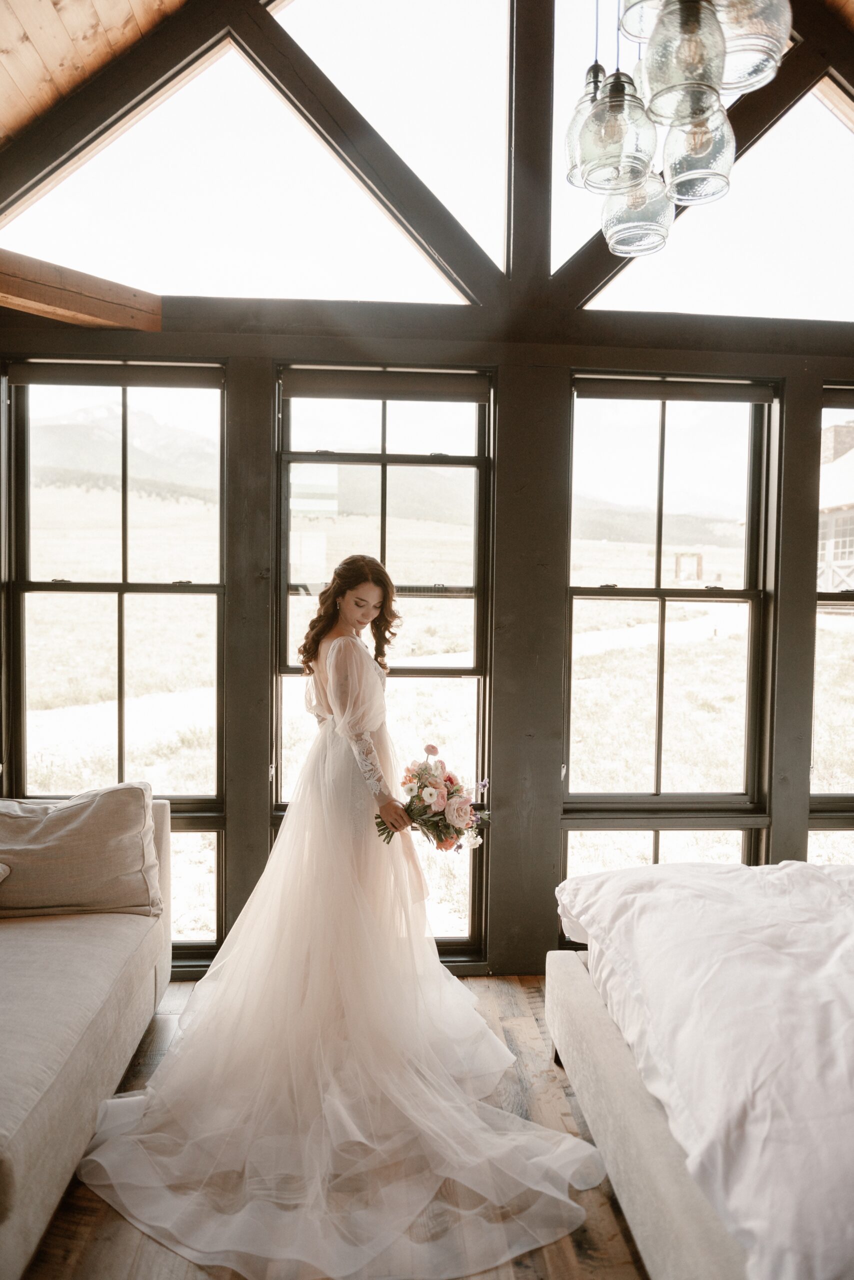 A photo of a bride posing in front of tall windows inside a cabin before her Three Peaks Ranch wedding. Photo by Colorado wedding photographer Ashley Joyce.