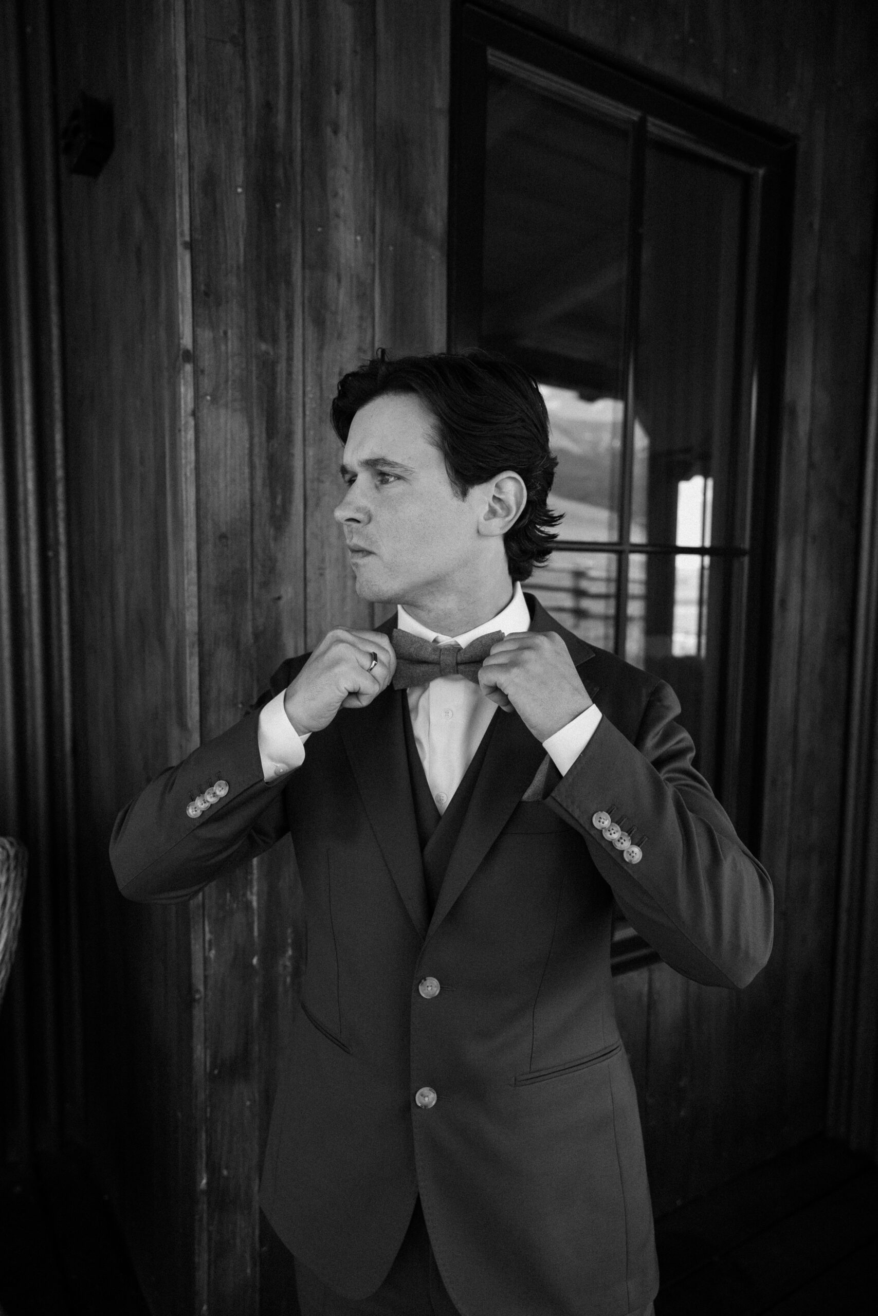 A black and white photo of groom, Bernard, holding on to his bowtie as he poses for his groom portraits at Three Peaks Ranch. Photo by Colorado wedding photographer Ashley Joyce.