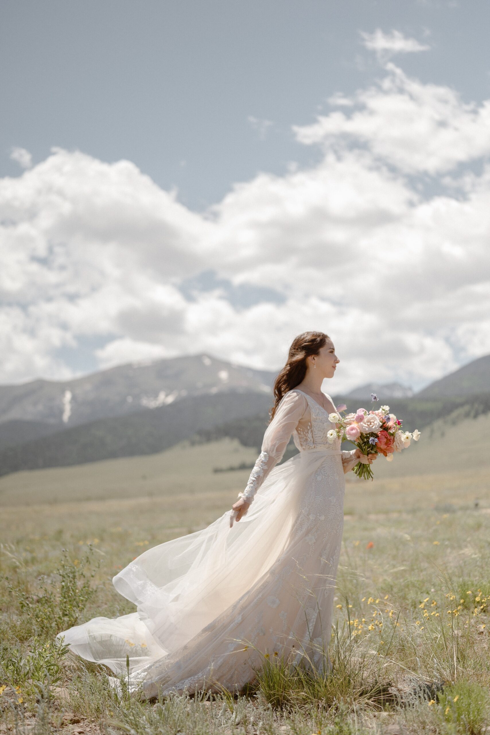 A bride stands in a grassy field while she holds her wedding bouquet as she poses for her bridal portraits at Three Peaks Ranch. Photo by Colorado wedding photographer Ashley Joyce.