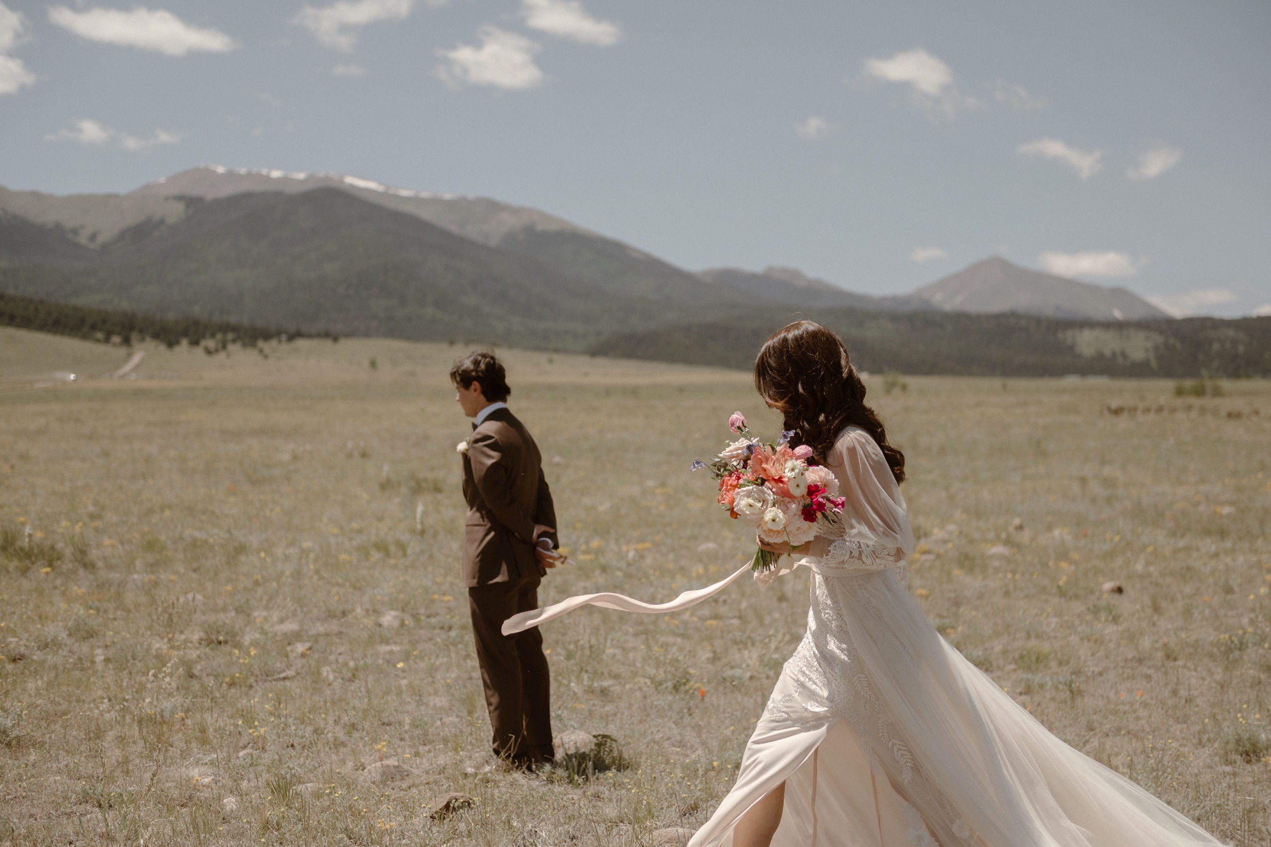 A bride walks through a grassy field to her fiance while he waits for her at Three Peaks Ranch. Photo by Colorado wedding photographer Ashley Joyce.