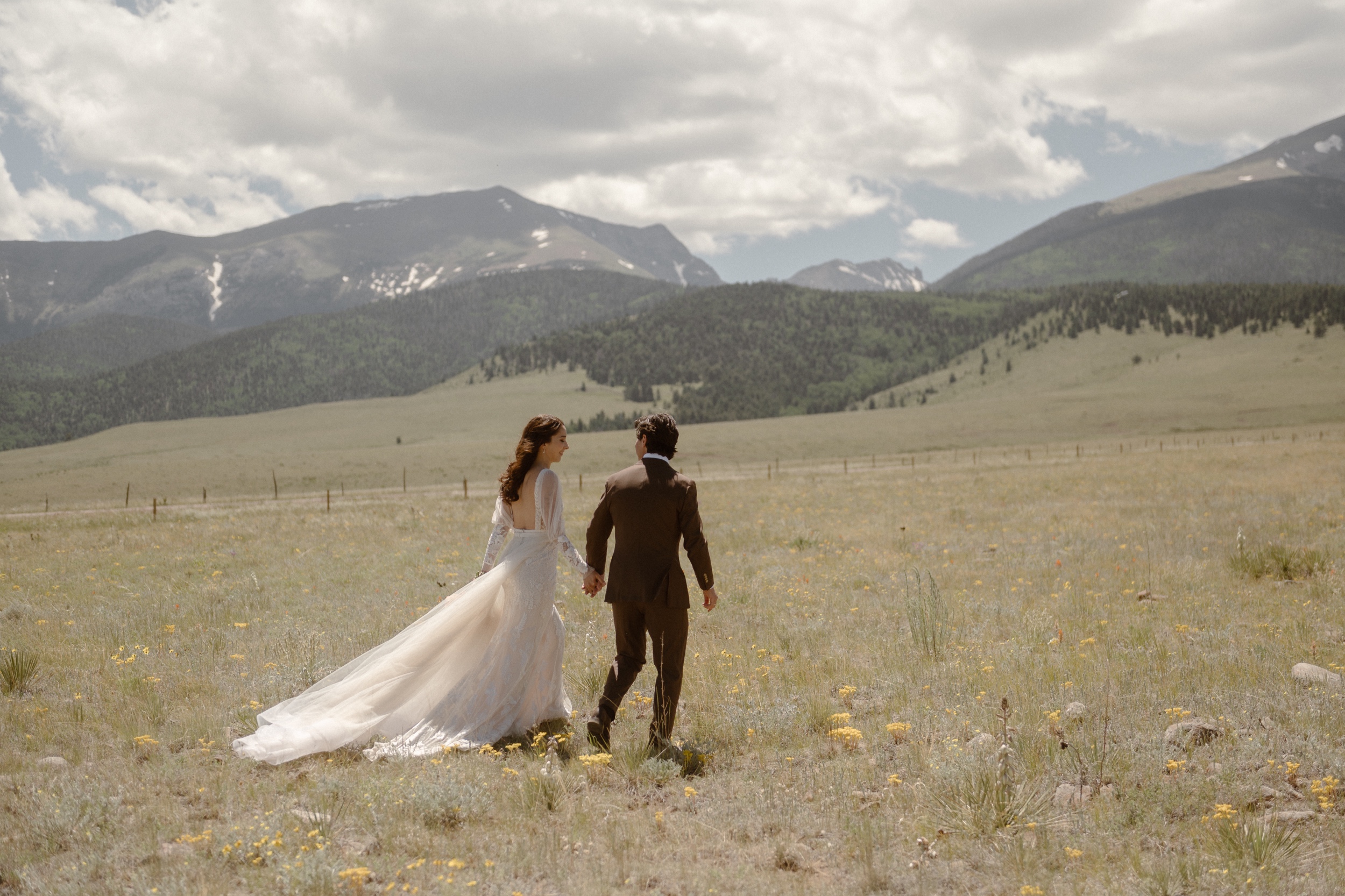 A bride and a groom walk away from the camera while they hold hands in a grassy field with mountains in the background. Photo by Colorado wedding photographer Ashley Joyce.
