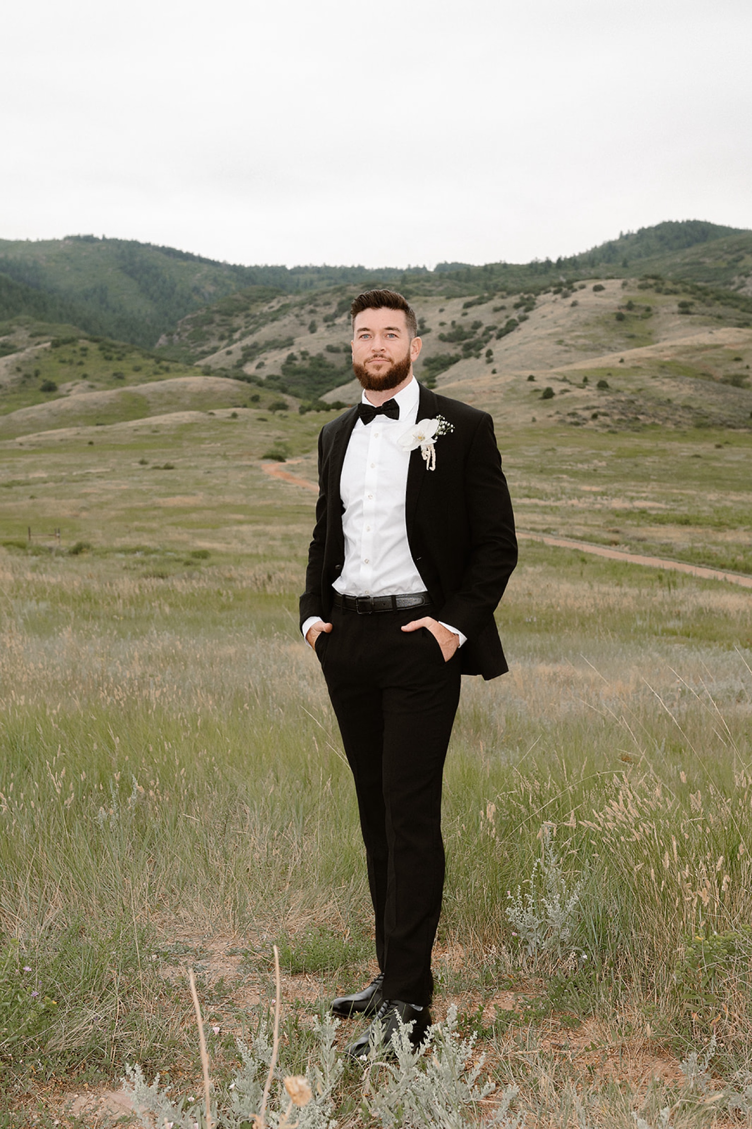 A flash photo of a groom posing for his wedding portraits in a field with mountains in the background