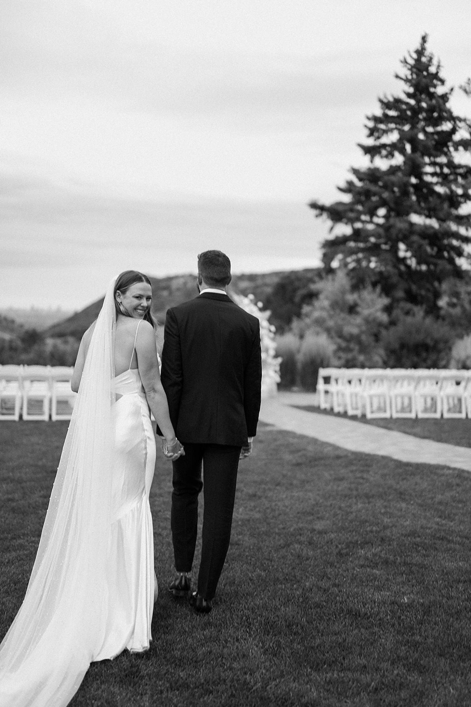 A bride and groom walk away from the camera while the bride turns back to smile at the camera