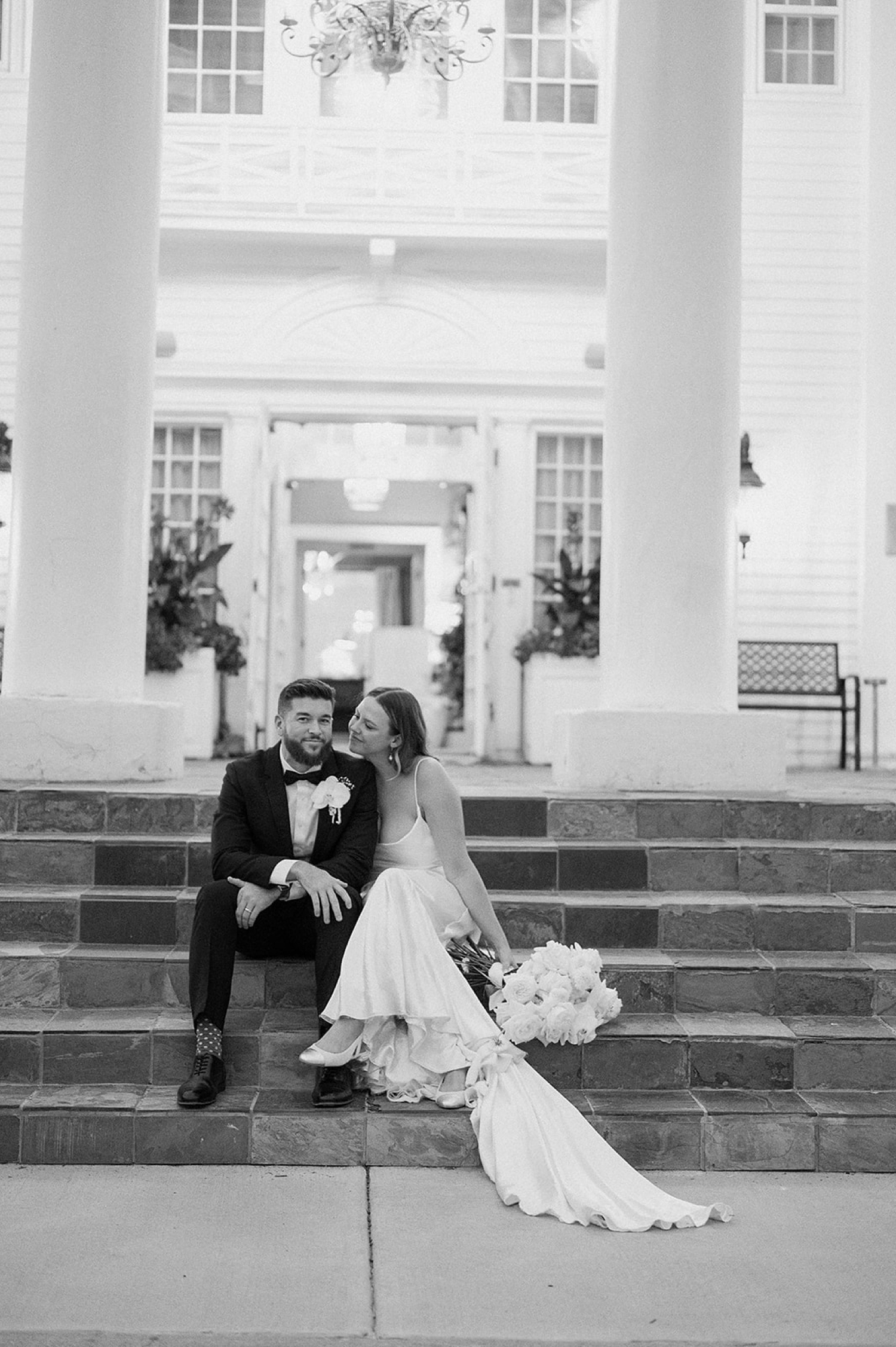 A bride and groom sit on the steps of The Manor House