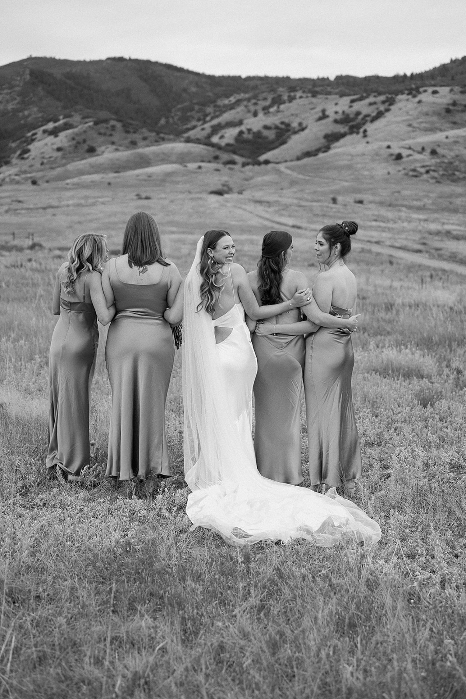 A bride hugs her bridesmaids from behind and turns to the camera to smile