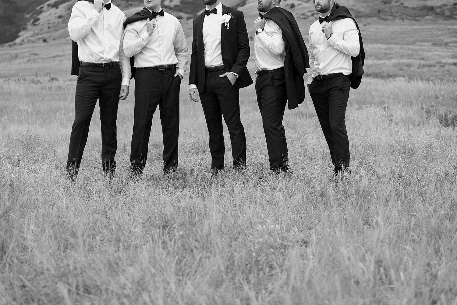 A groom and his groomsmen stand in a field of grass while they hold their suit jackets over their shoulders