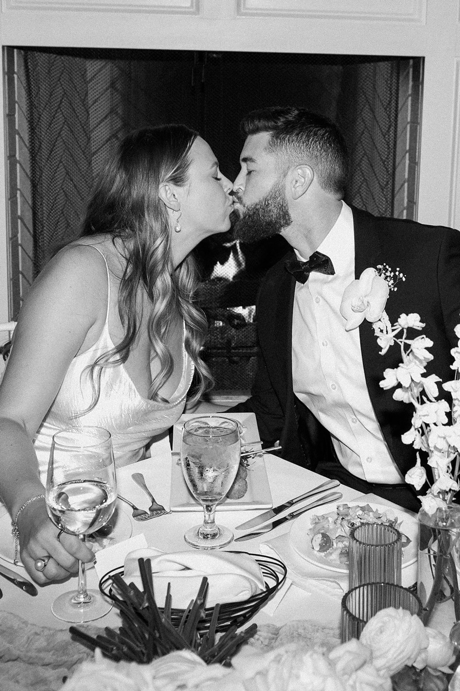 A bride and groom sit at their wedding reception table and kiss one another