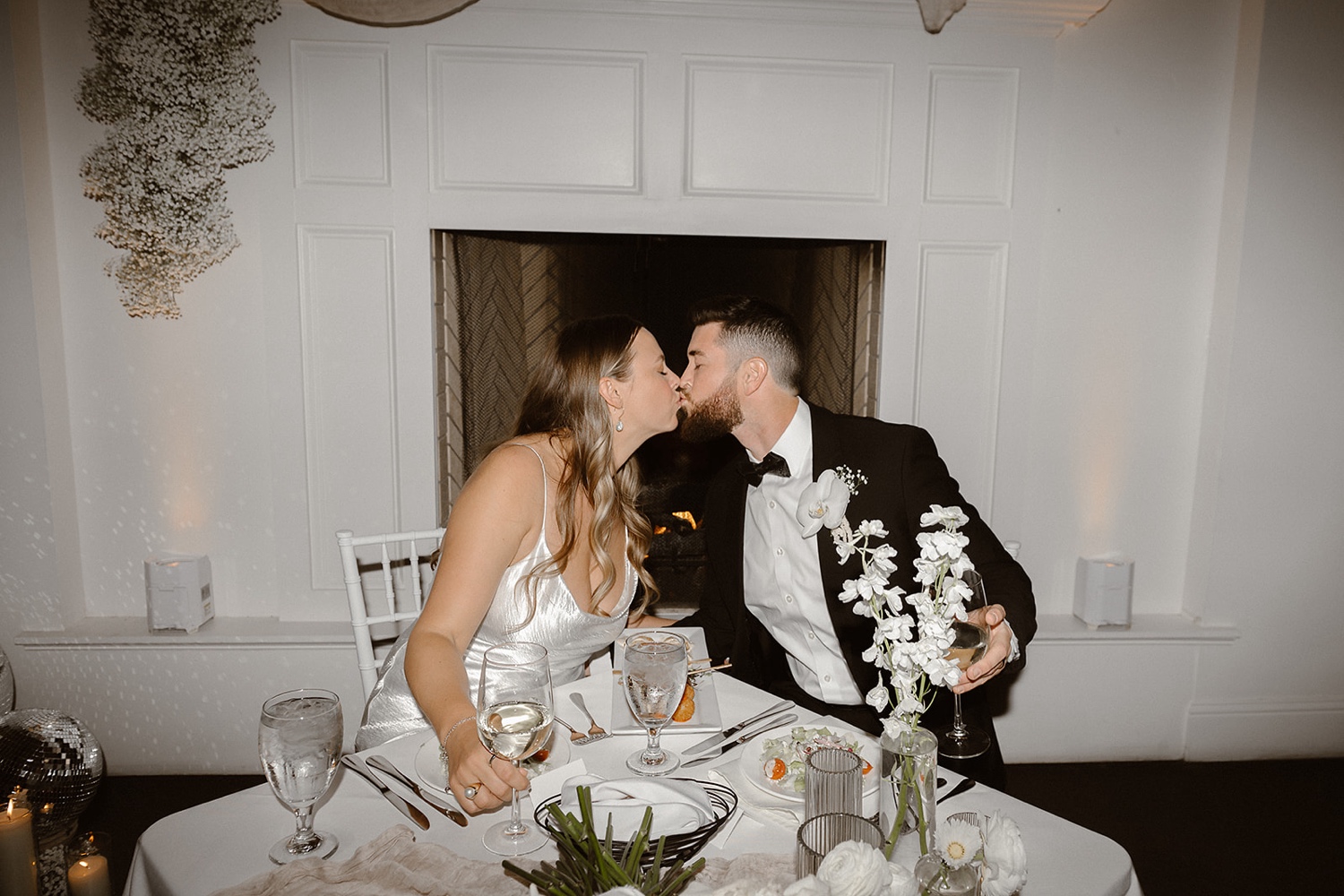 A bride and groom sit at their wedding reception table and kiss one another