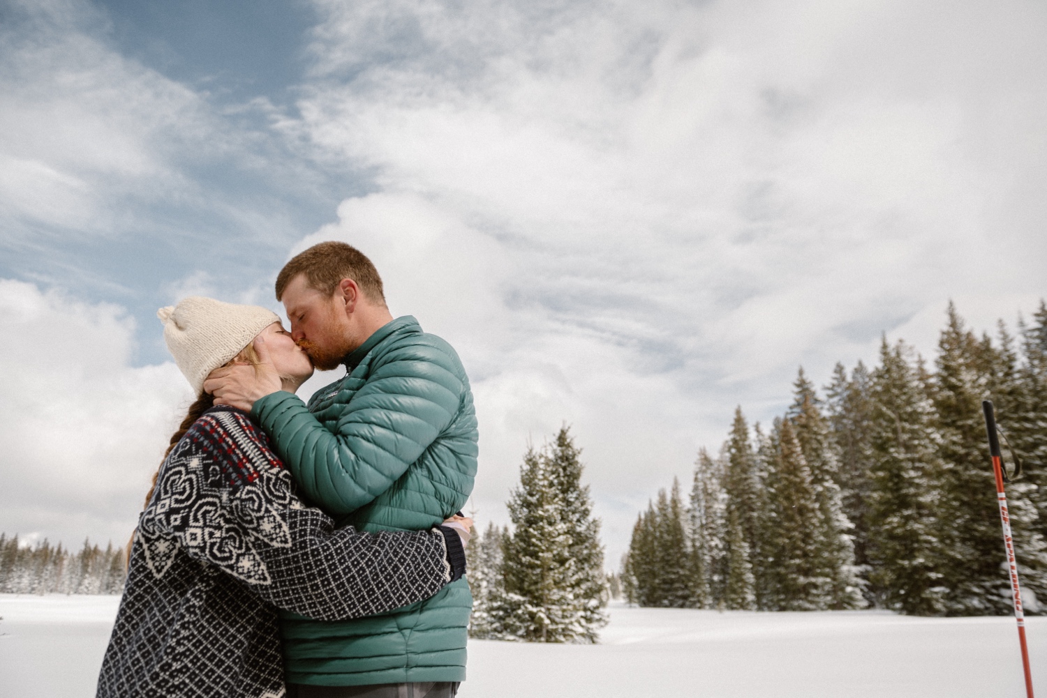 Couple stops for a photo break while cross-country skiing, with snow-covered mountains and winter engagement outfits in the background.