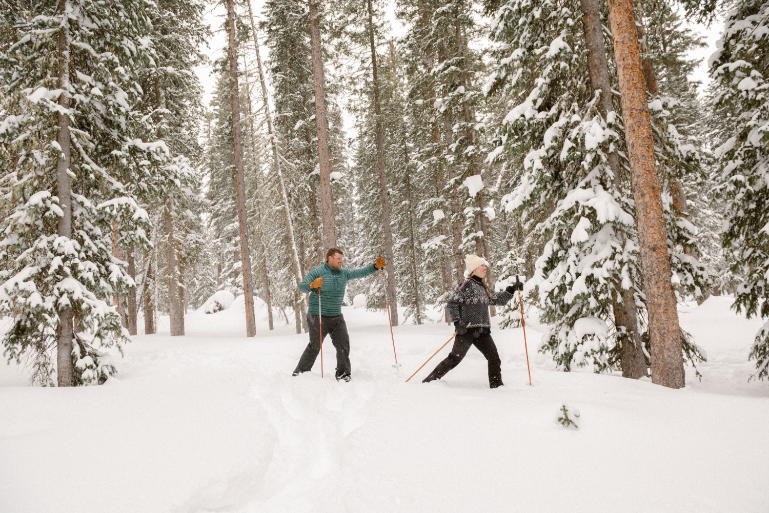 Couple cross-country skiing in snowy forest, wearing winter engagement outfits, during their winter engagement session near Montrose, Colorado.