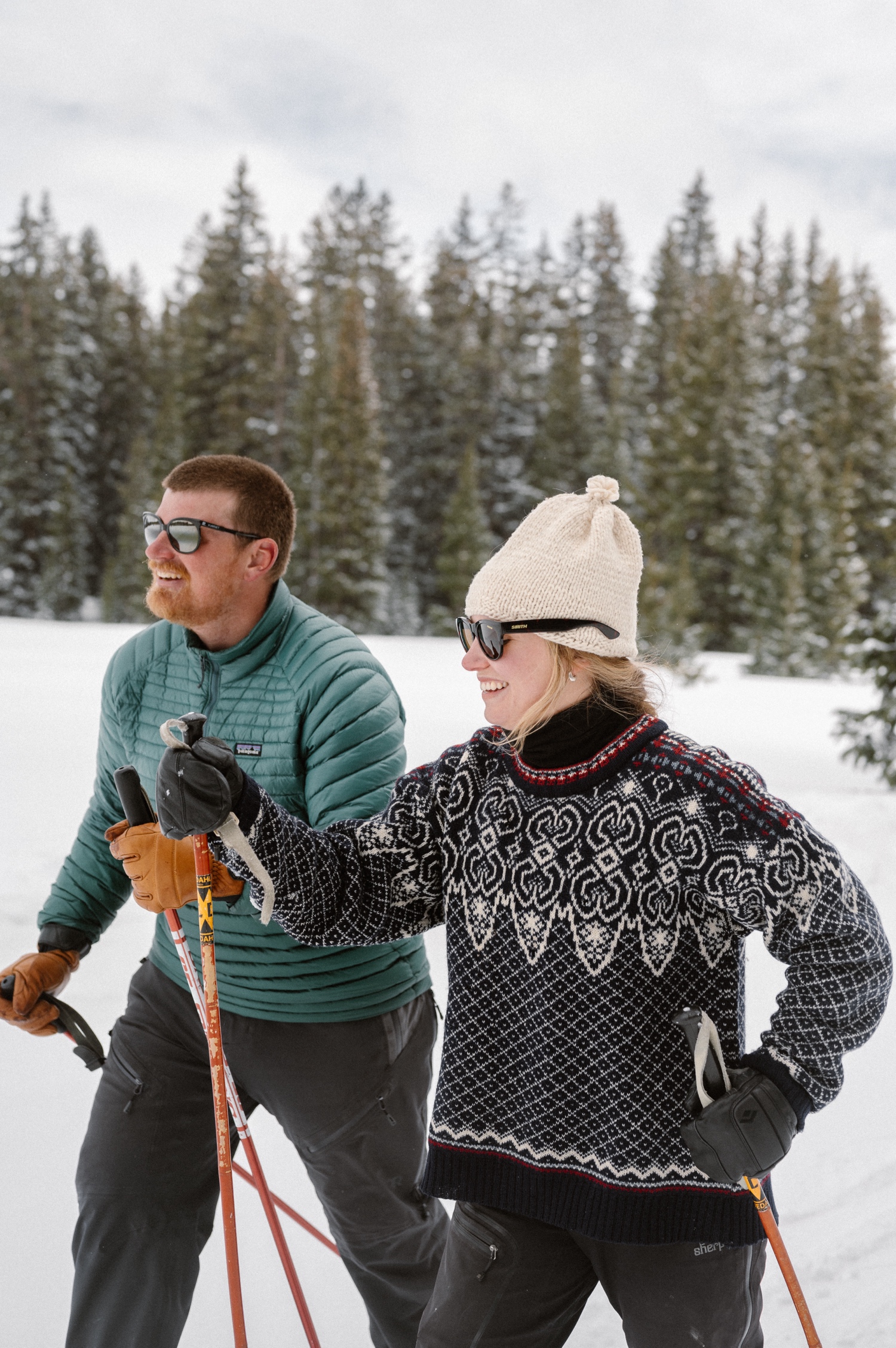 A couple laughing together while bundled up in cozy coats during their winter engagement photo session on cross-country skis in Montrose, Colorado.