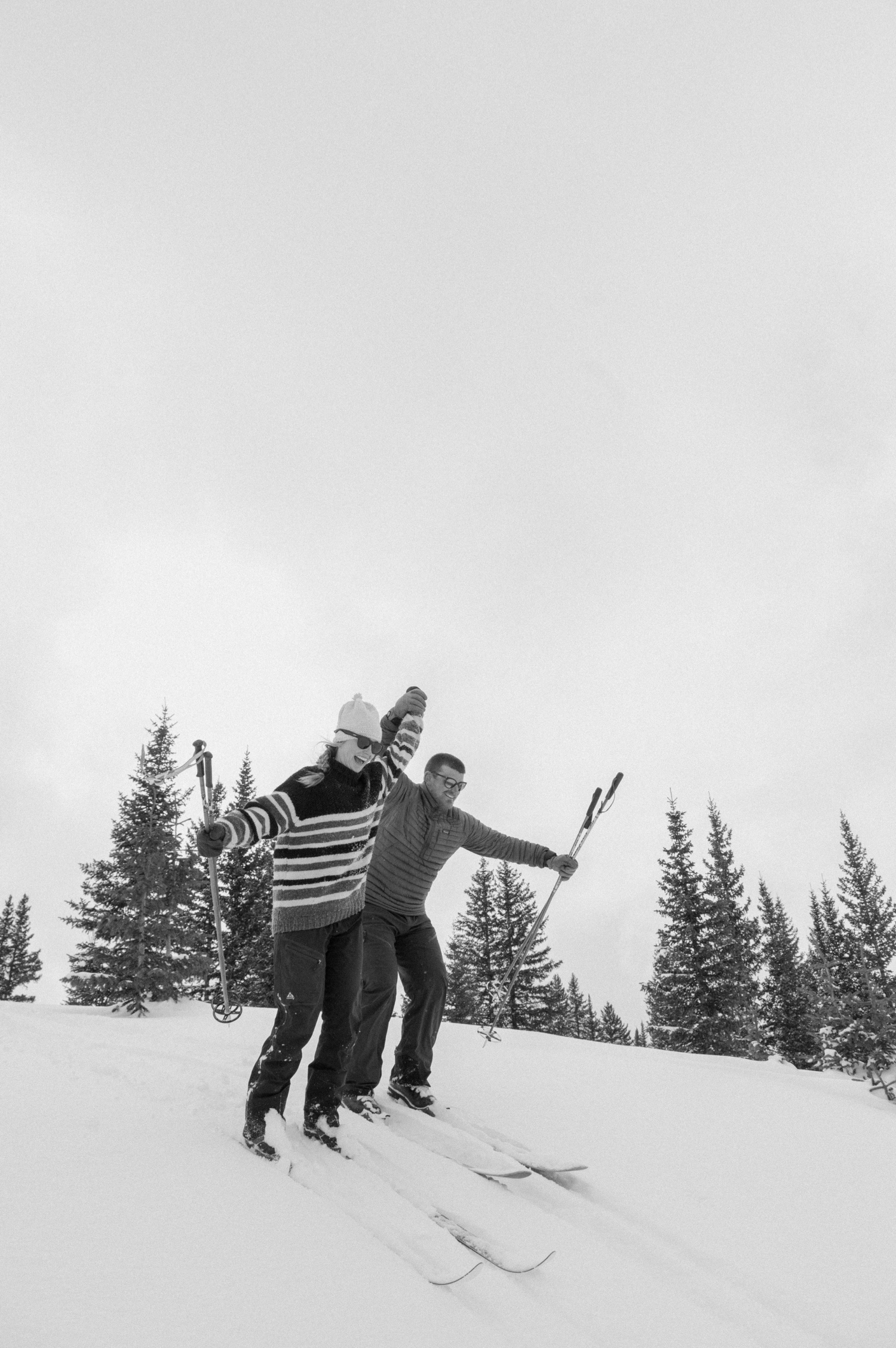 Couple cross-country skiing in snowy forest, wearing winter engagement outfits, during their winter engagement session near Montrose, Colorado.