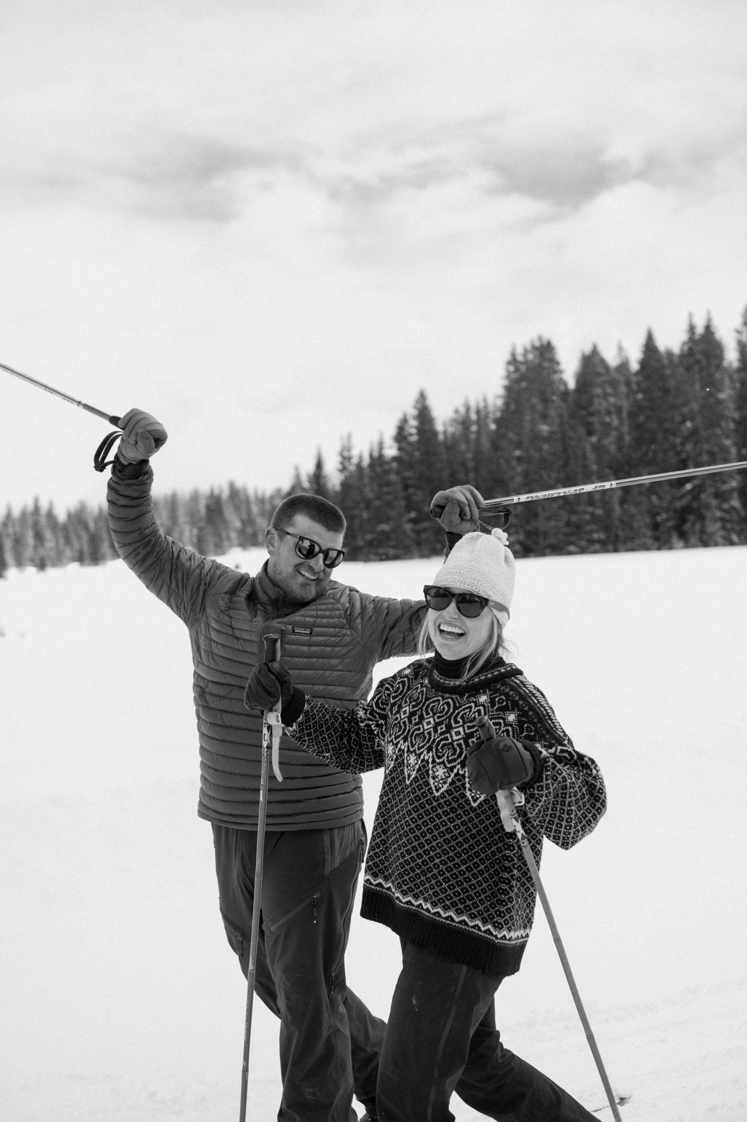 Couple cross-country skiing in snowy forest, wearing winter engagement outfits, during their winter engagement session near Montrose, Colorado.