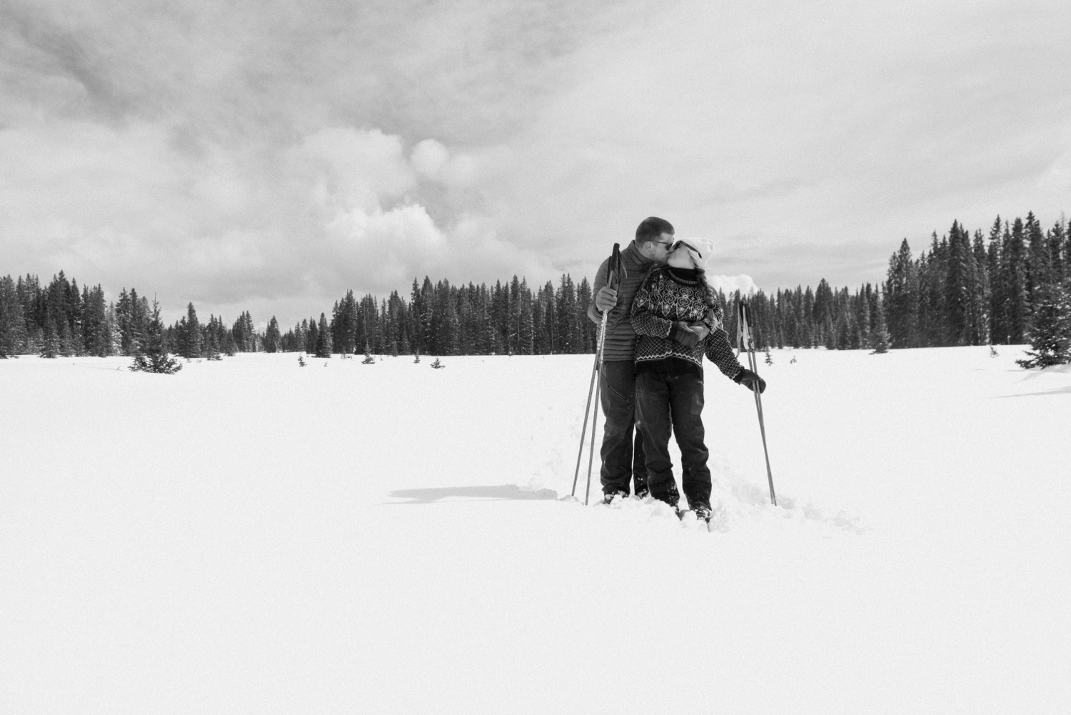 Couple cross-country skiing in snowy forest, wearing winter engagement outfits, during their winter engagement session near Montrose, Colorado.